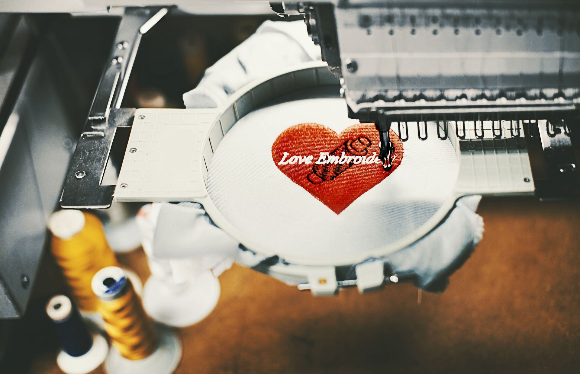 Person sewing an embroidered flower design on denim, holding needle. Wooden table with scattered supplies.