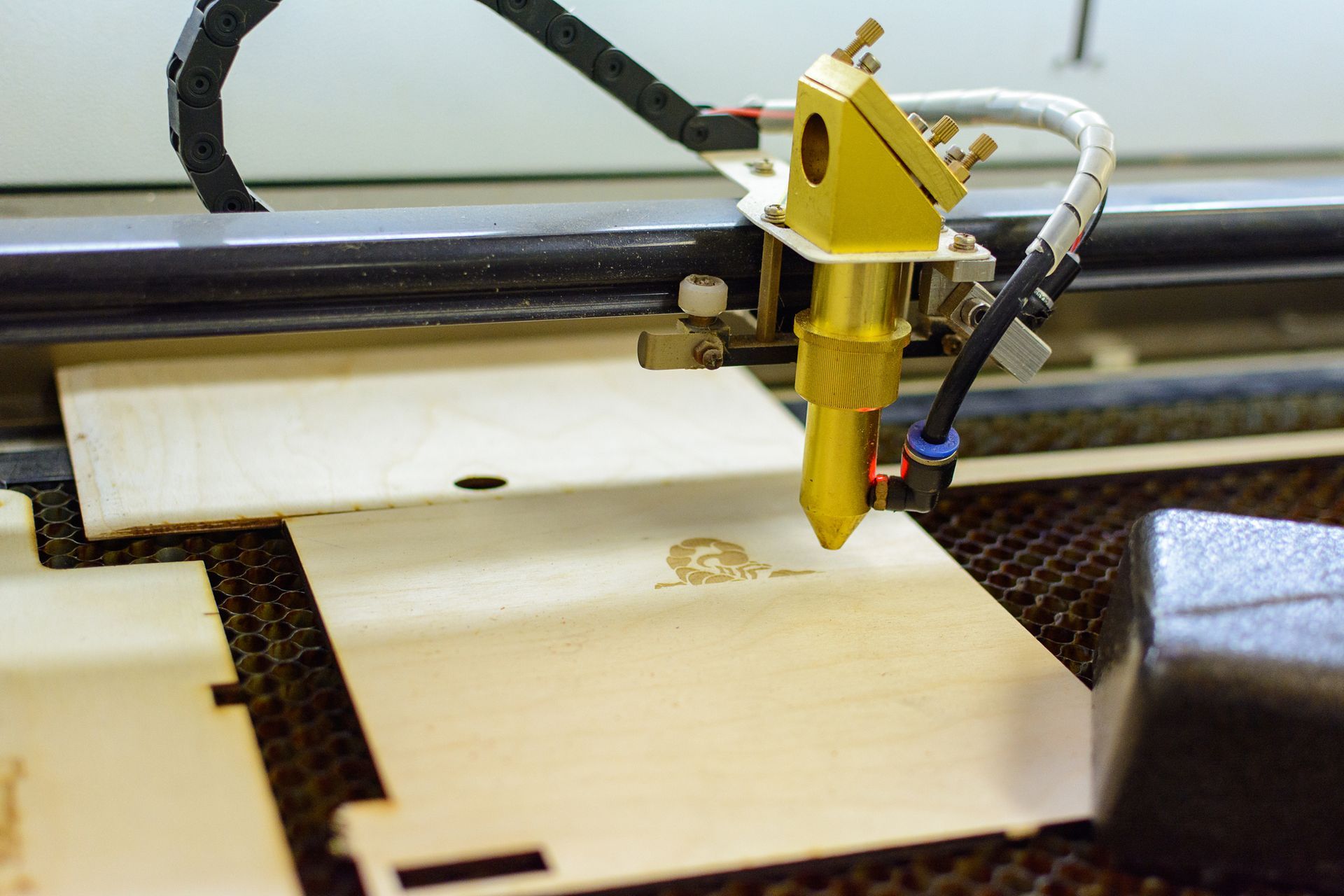 Laser cutter engraving a design onto a light-colored wooden sheet. The laser head is gold, and the machine is in operation.