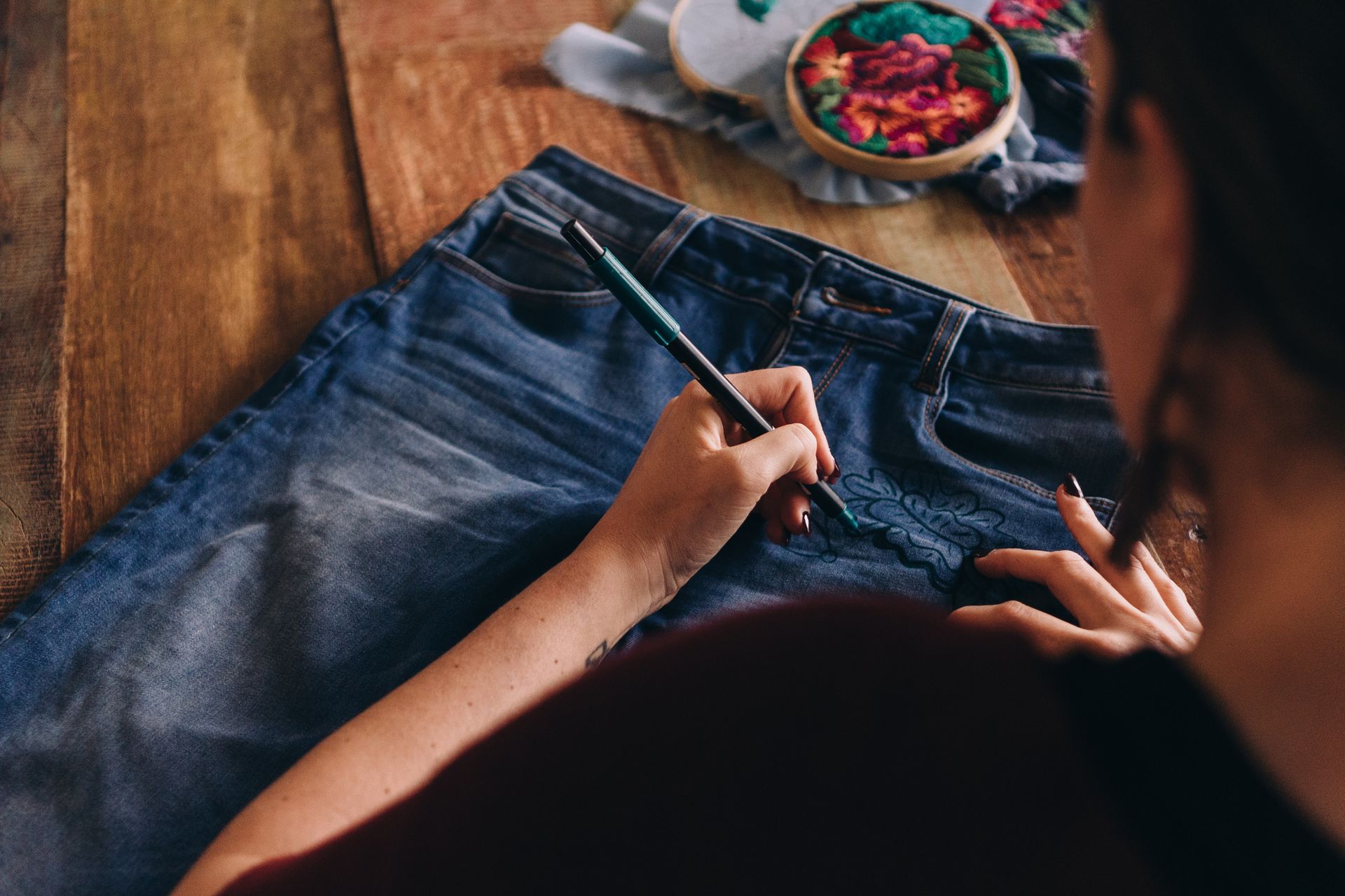 Person painting on a pair of blue jeans with a paintbrush, embroidery hoop in background.
