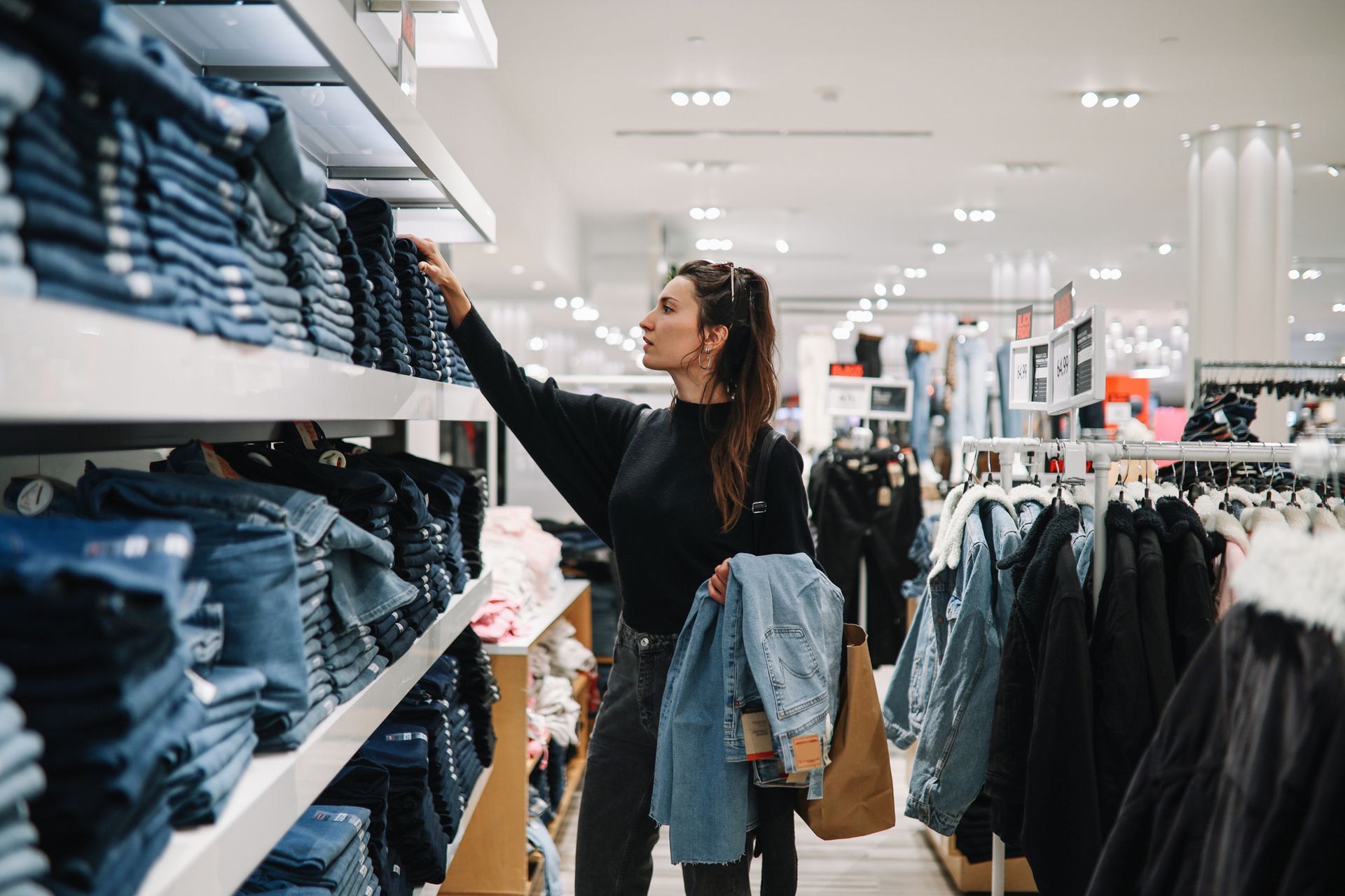 Woman shopping in a clothing store, reaching for a stack of blue jeans on a shelf.