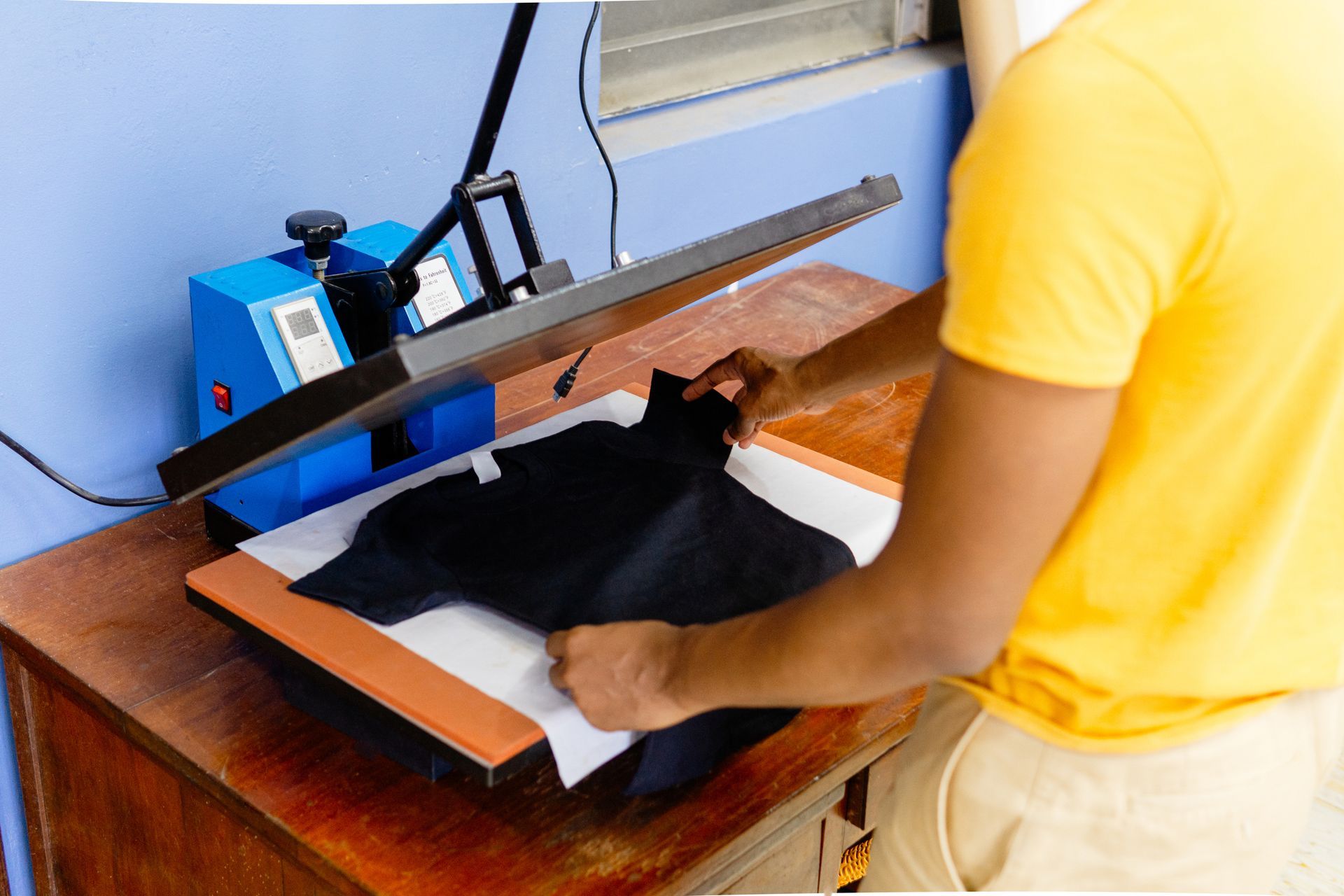 Person placing a black shirt on a heat press machine on a wooden table. Yellow shirt, blue wall.