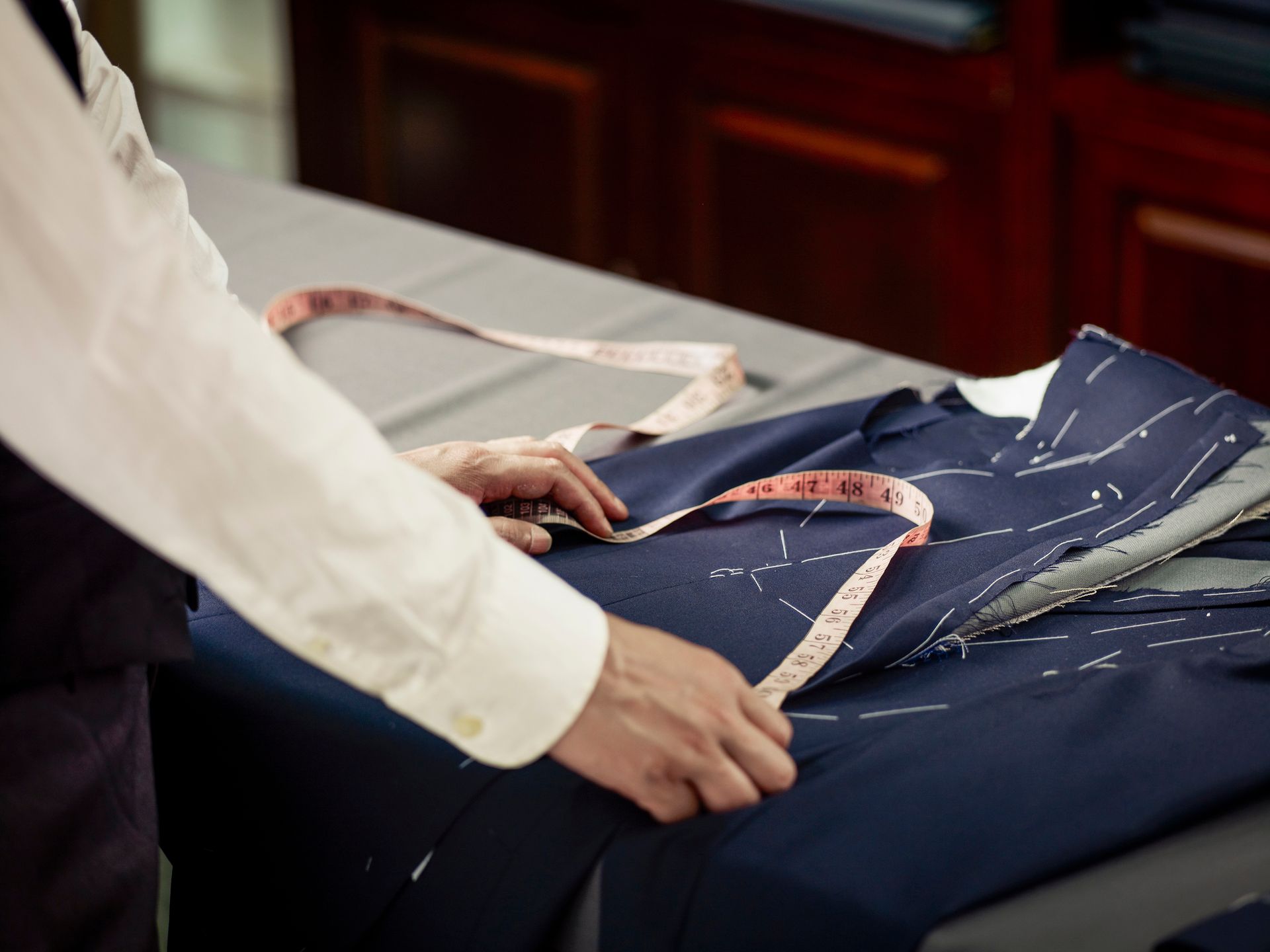 Tailor measuring a dark blue suit jacket with a pink measuring tape on a table.