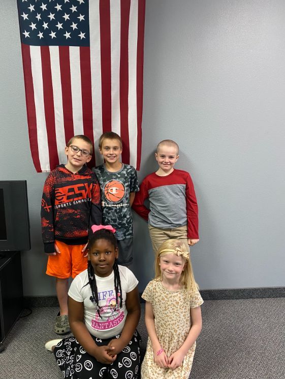 Children sitting on chairs in a room, smiling and waving. One child wears sunglasses. Neutral setting.
