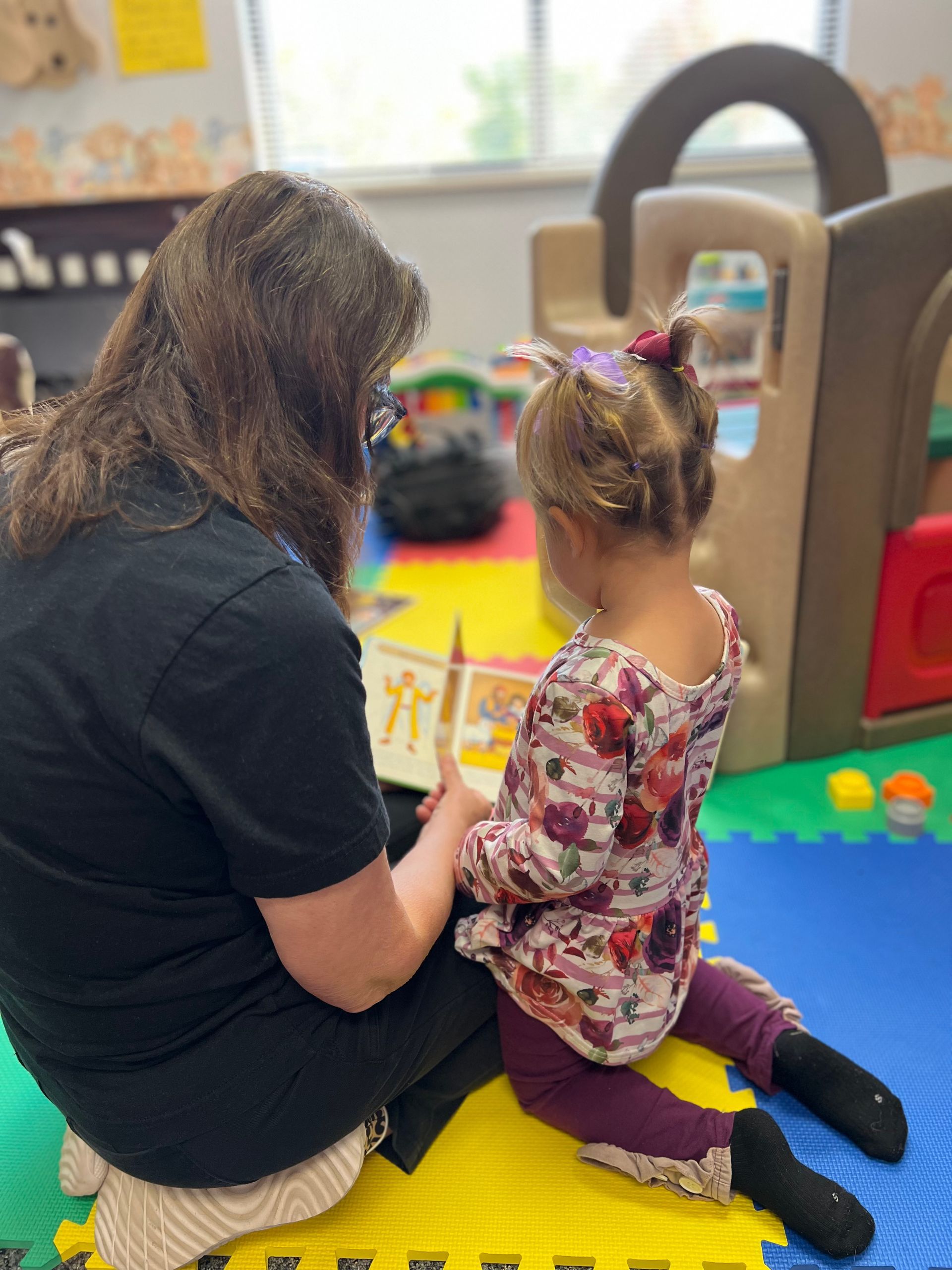 Woman with two children in a colorful room smiles. Children point; toys and giraffe decor.