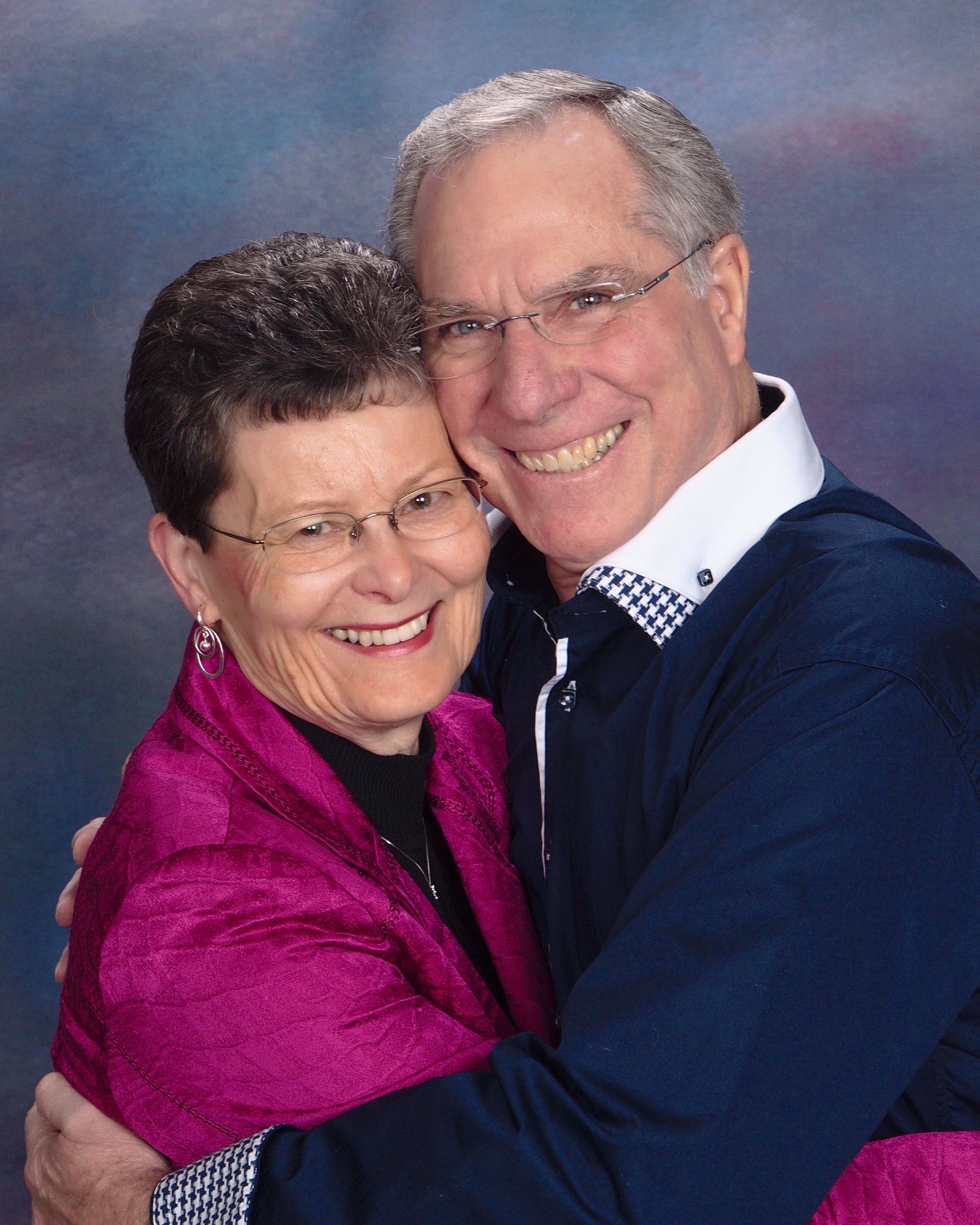 Smiling couple embracing, man in glasses hugs woman wearing pink wrap, blue background.