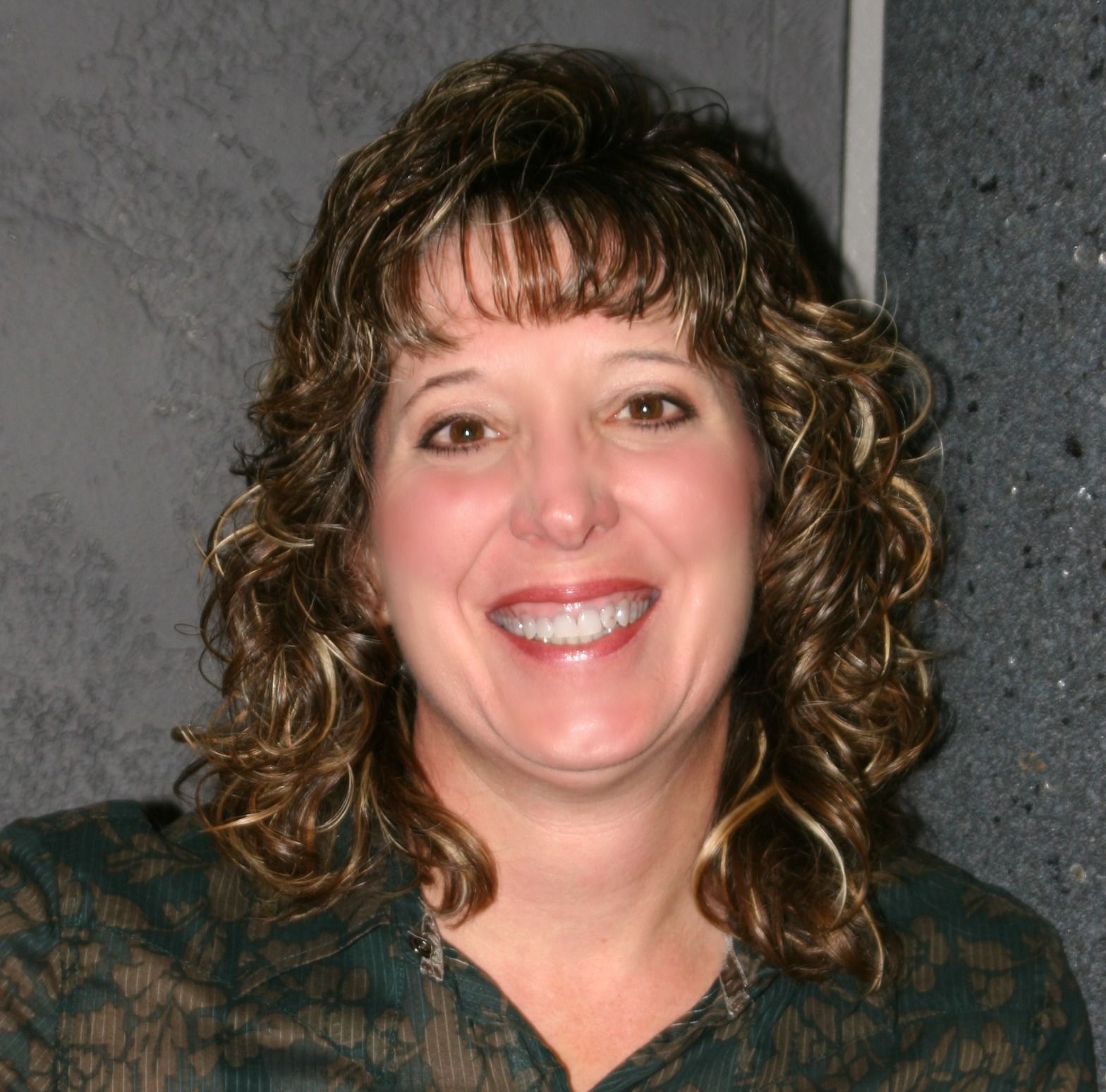 Woman with curly brown hair smiling, wearing a dark green shirt, in front of a gray wall.