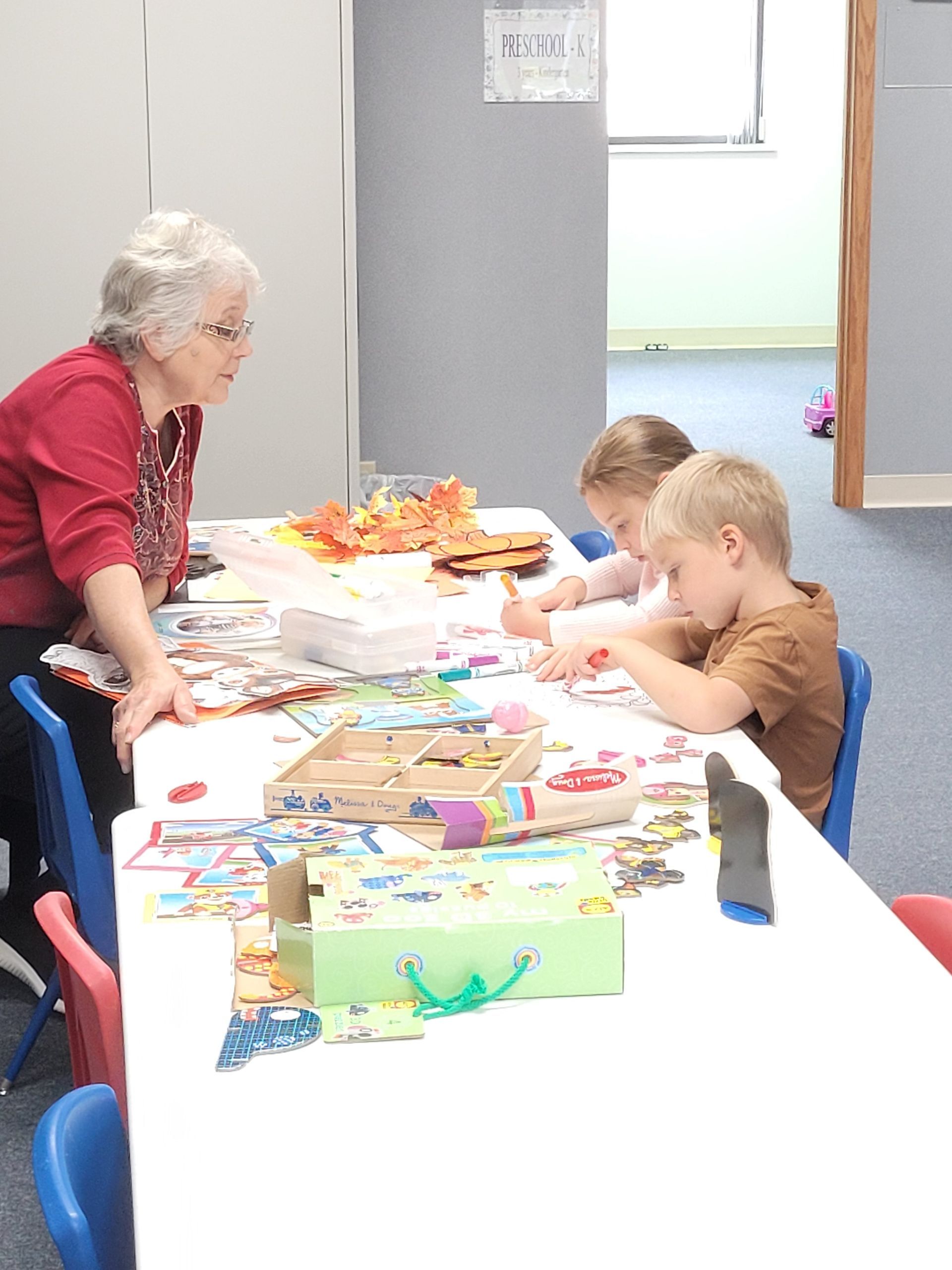 Woman assisting two children at a table covered in toys and craft supplies.