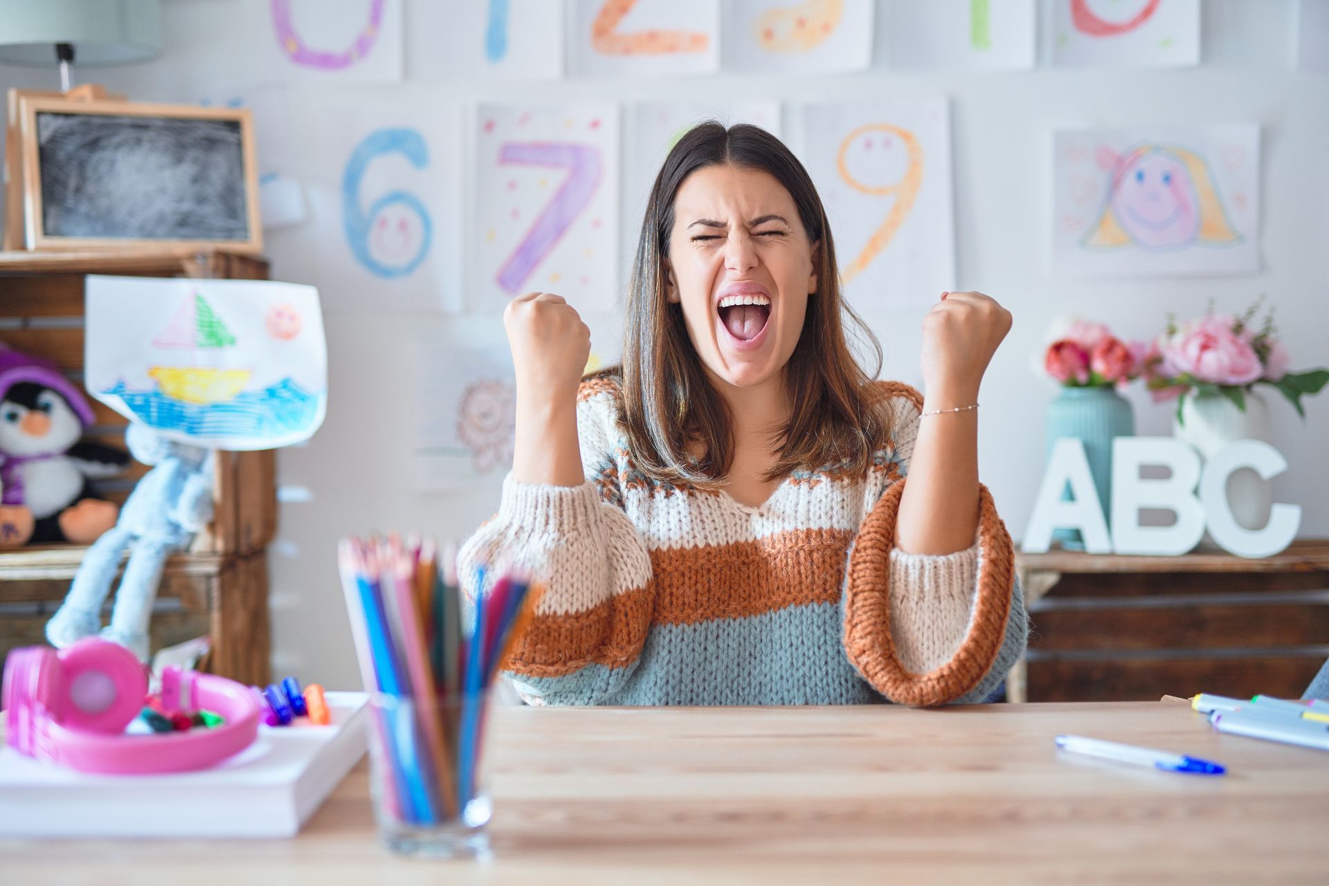 Woman cheering at a colorful desk, with art supplies and ABC blocks in the background.
