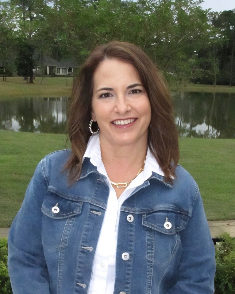 Woman in a denim jacket smiles outdoors near a pond.