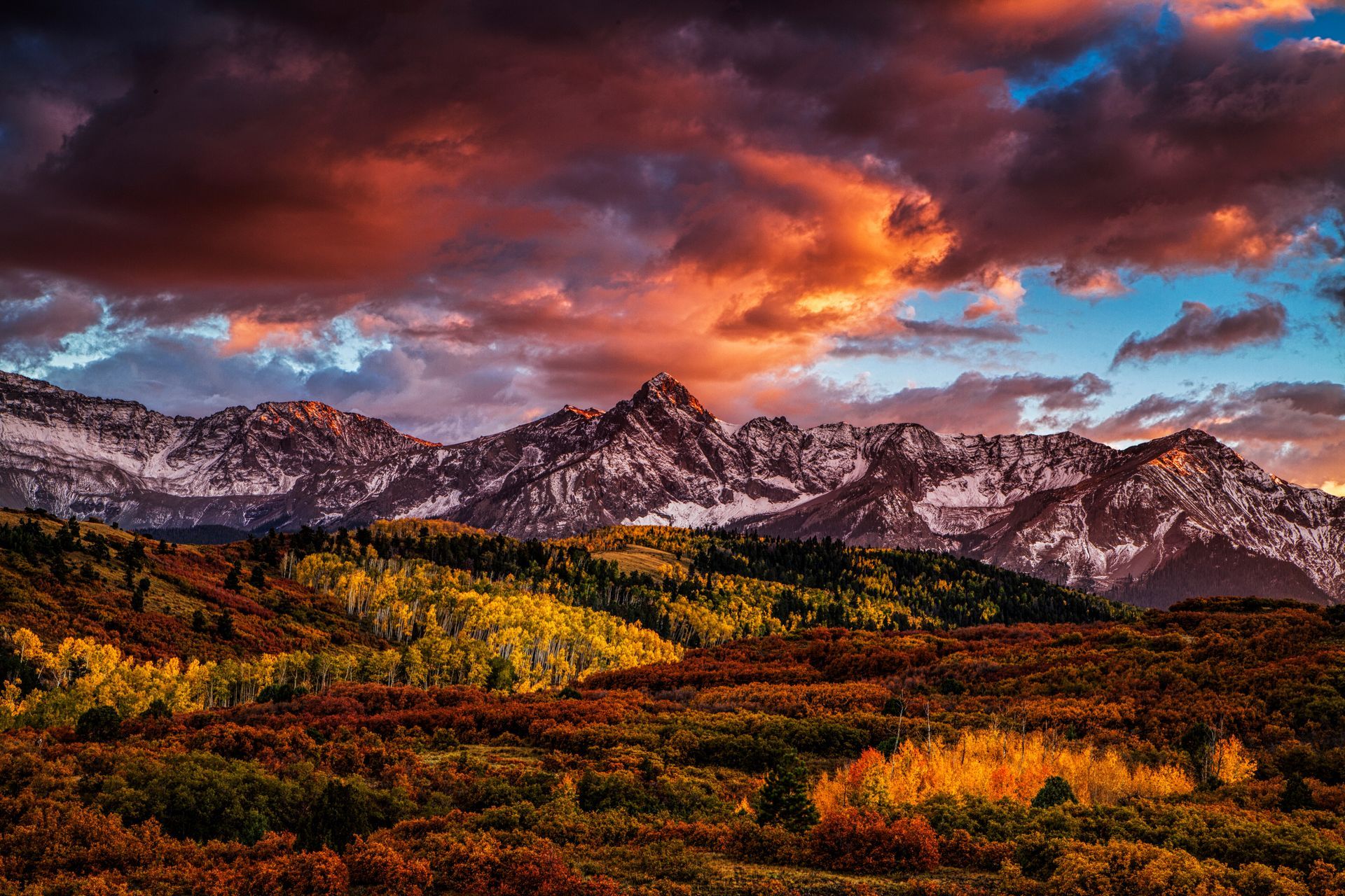 A sunset over a mountain range with trees in the foreground and a cloudy sky in the background.