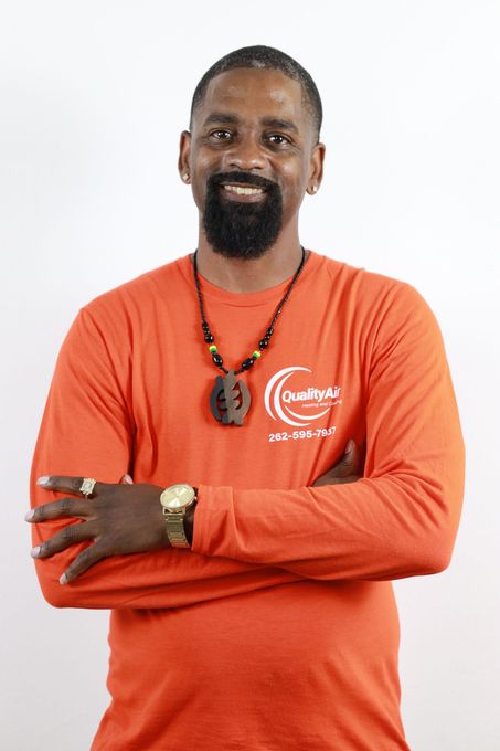 Man in orange shirt with arms crossed, smiling, wearing jewelry against white background.