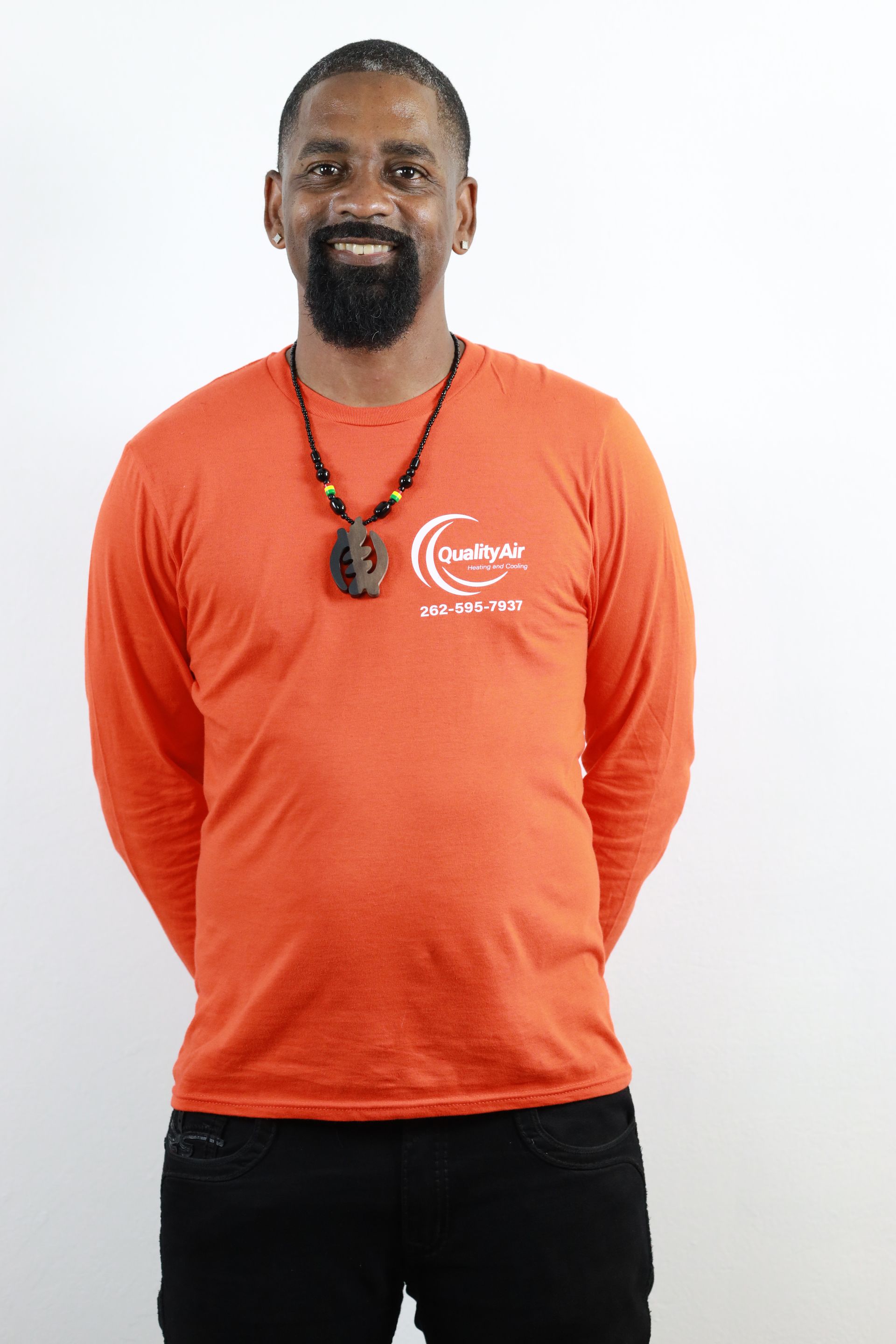 Man with a beard, wearing an orange long-sleeved shirt with a logo and black pants, standing in front of a white wall.