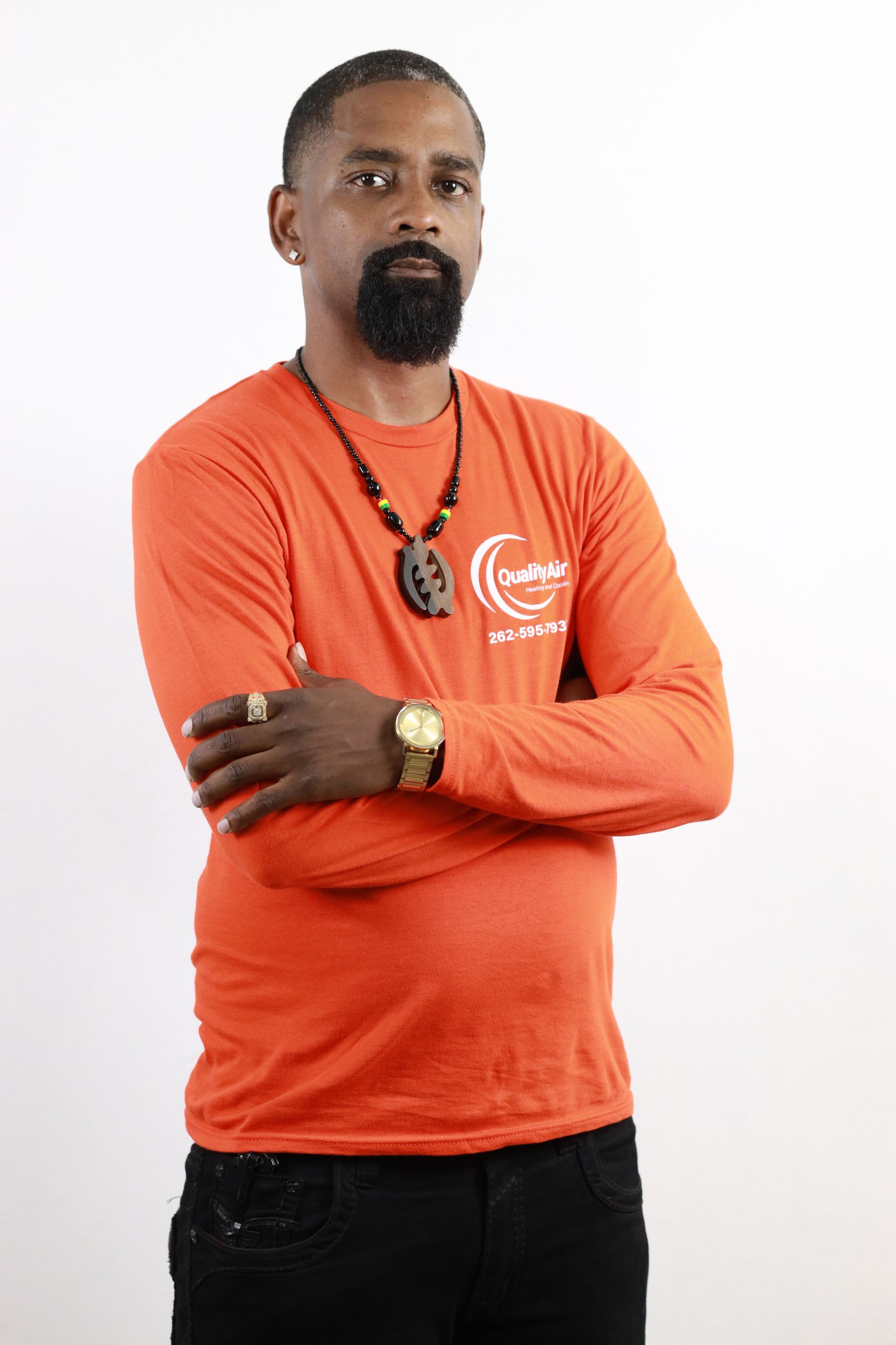 Man with beard wearing orange long-sleeved shirt, arms crossed, jewelry. Plain white background.