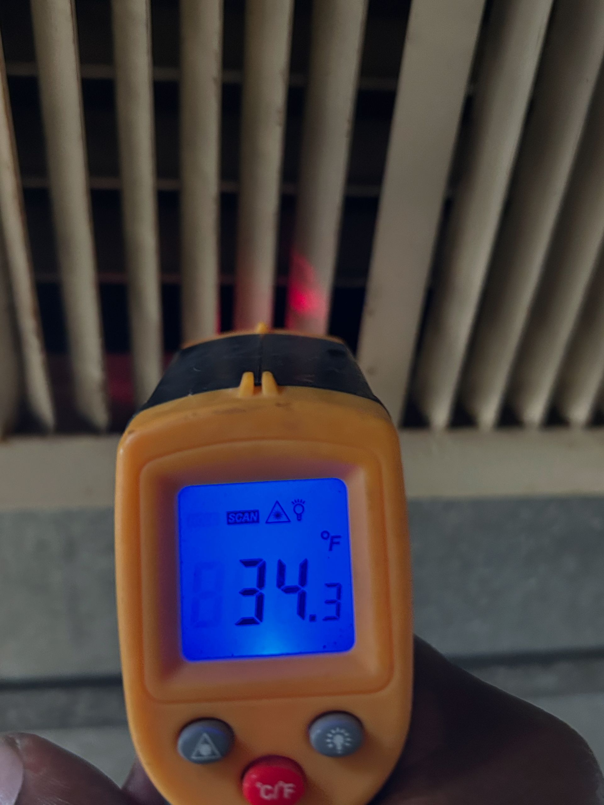 A handheld infrared thermometer measures 34.3 degrees Fahrenheit on an overhead vent, with a red laser sight.