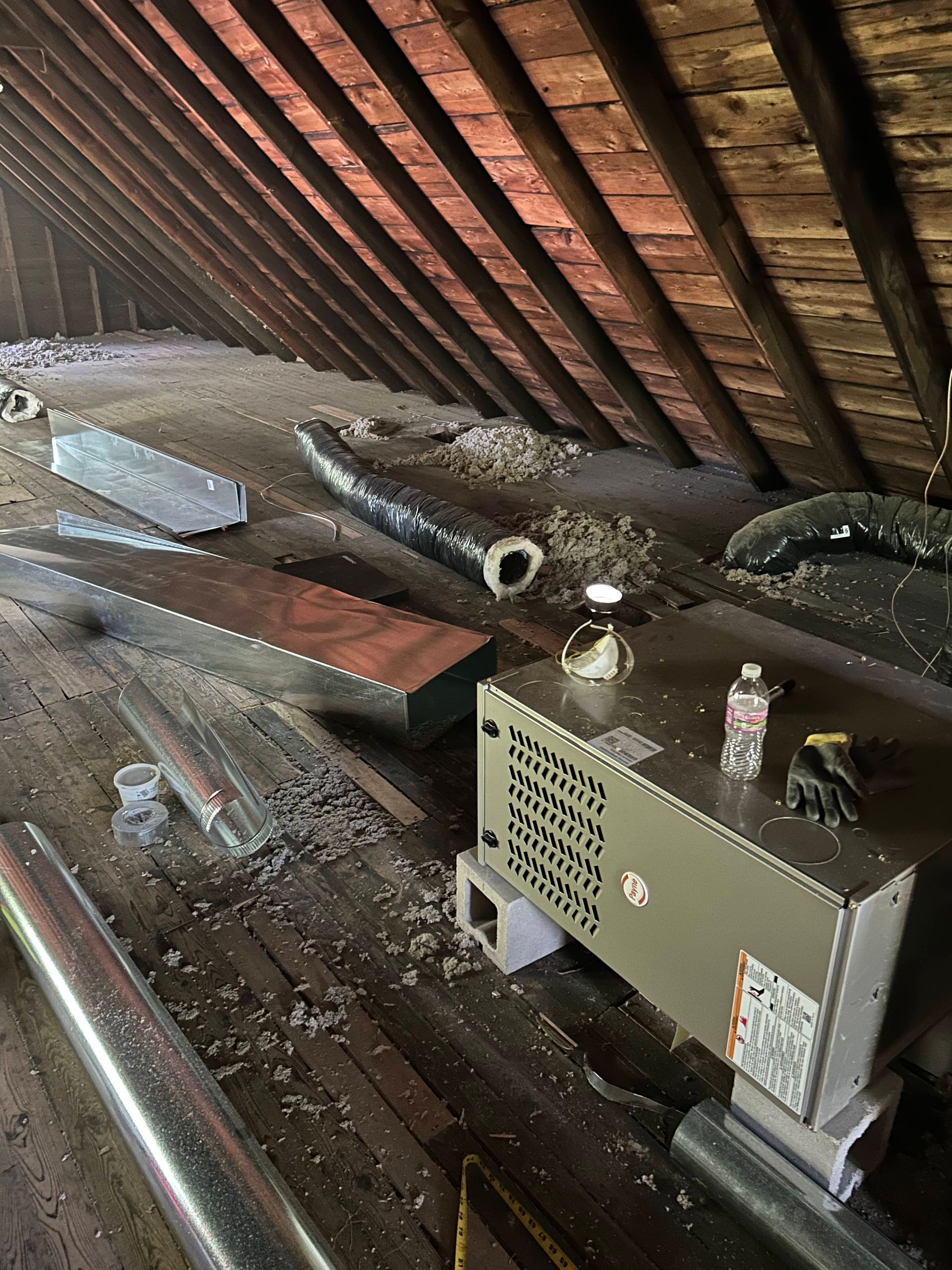 HVAC unit in an attic with ductwork and building materials scattered on wooden beams.