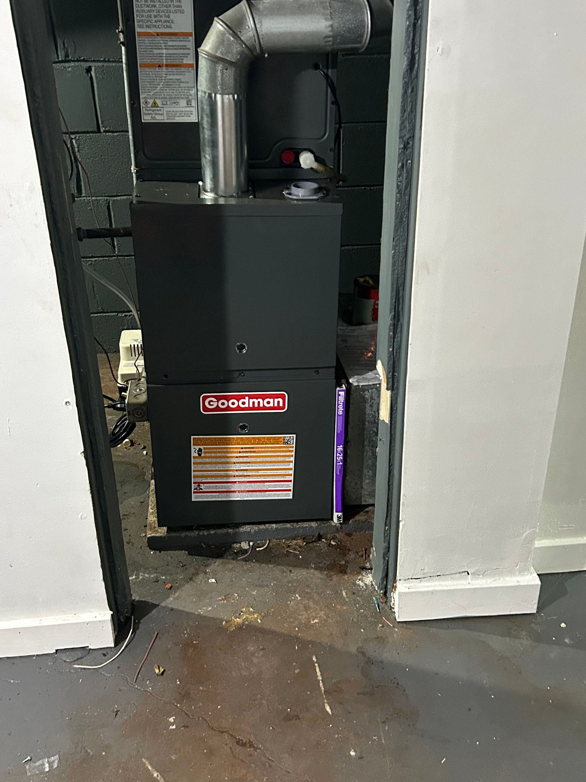 A Goodman furnace installed in a room. The unit is black with a silver exhaust pipe. The room has white walls and a gray floor.