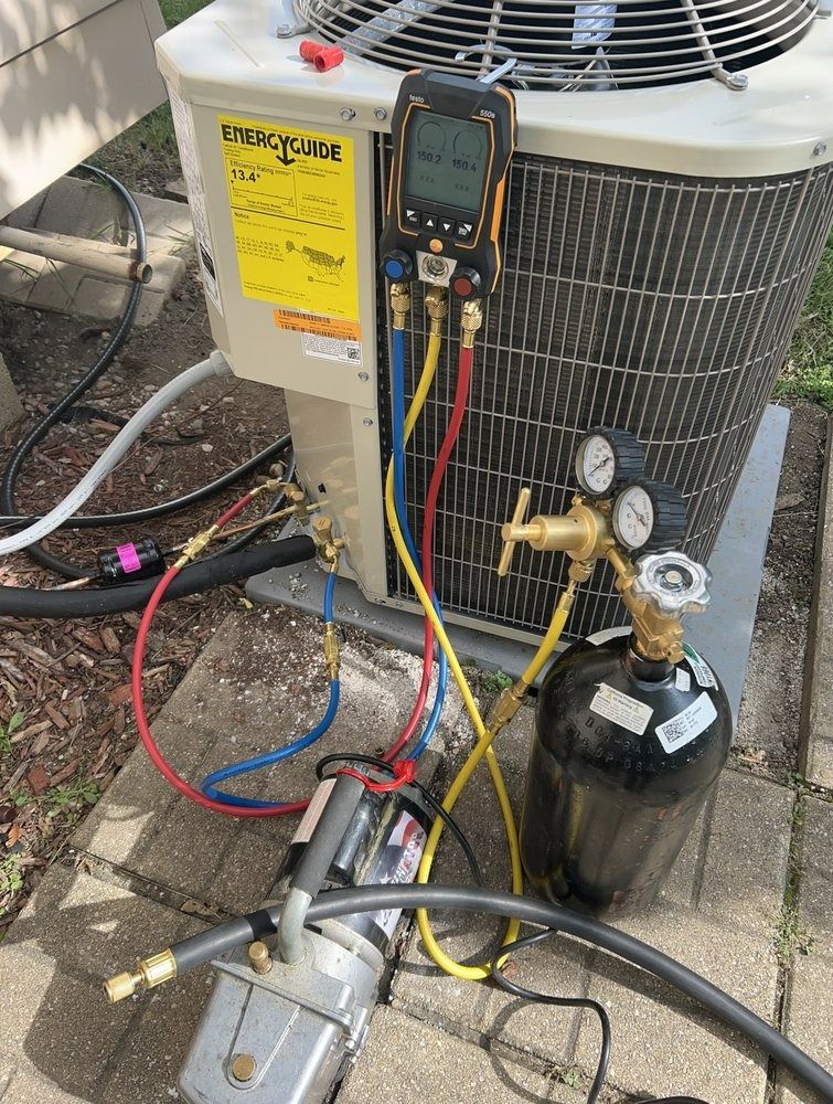 HVAC technician servicing an air conditioning unit. Equipment includes gauges, hoses, and a vacuum pump, outdoors on a patio.
