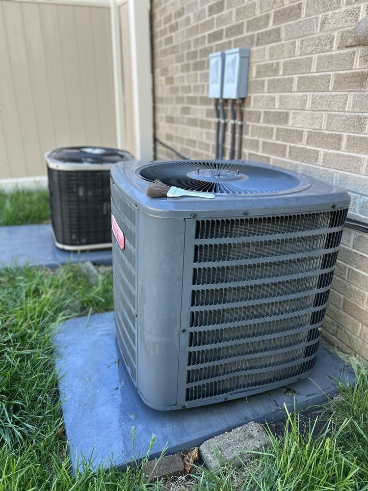 Two air conditioning units sit on concrete pads next to a brick building and a white fence.