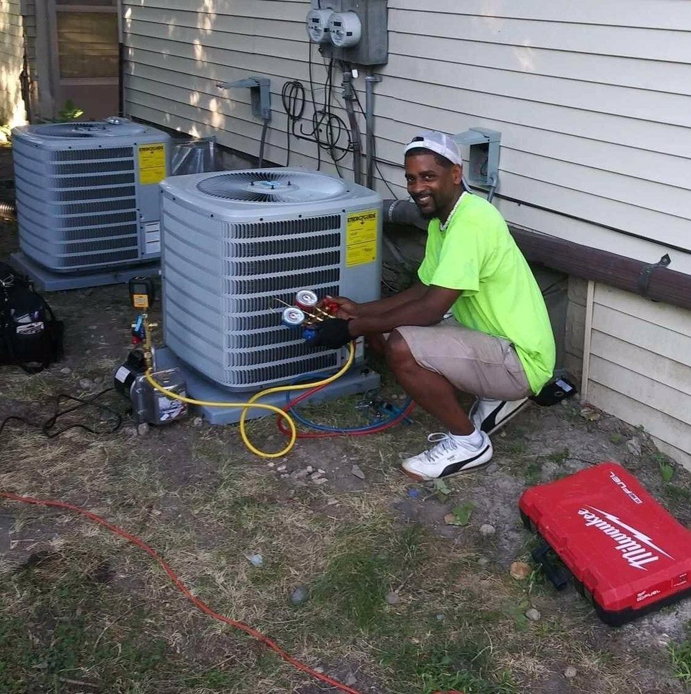 HVAC technician checks AC unit. Man in neon shirt kneels outside. Units, hoses, and toolbox visible.