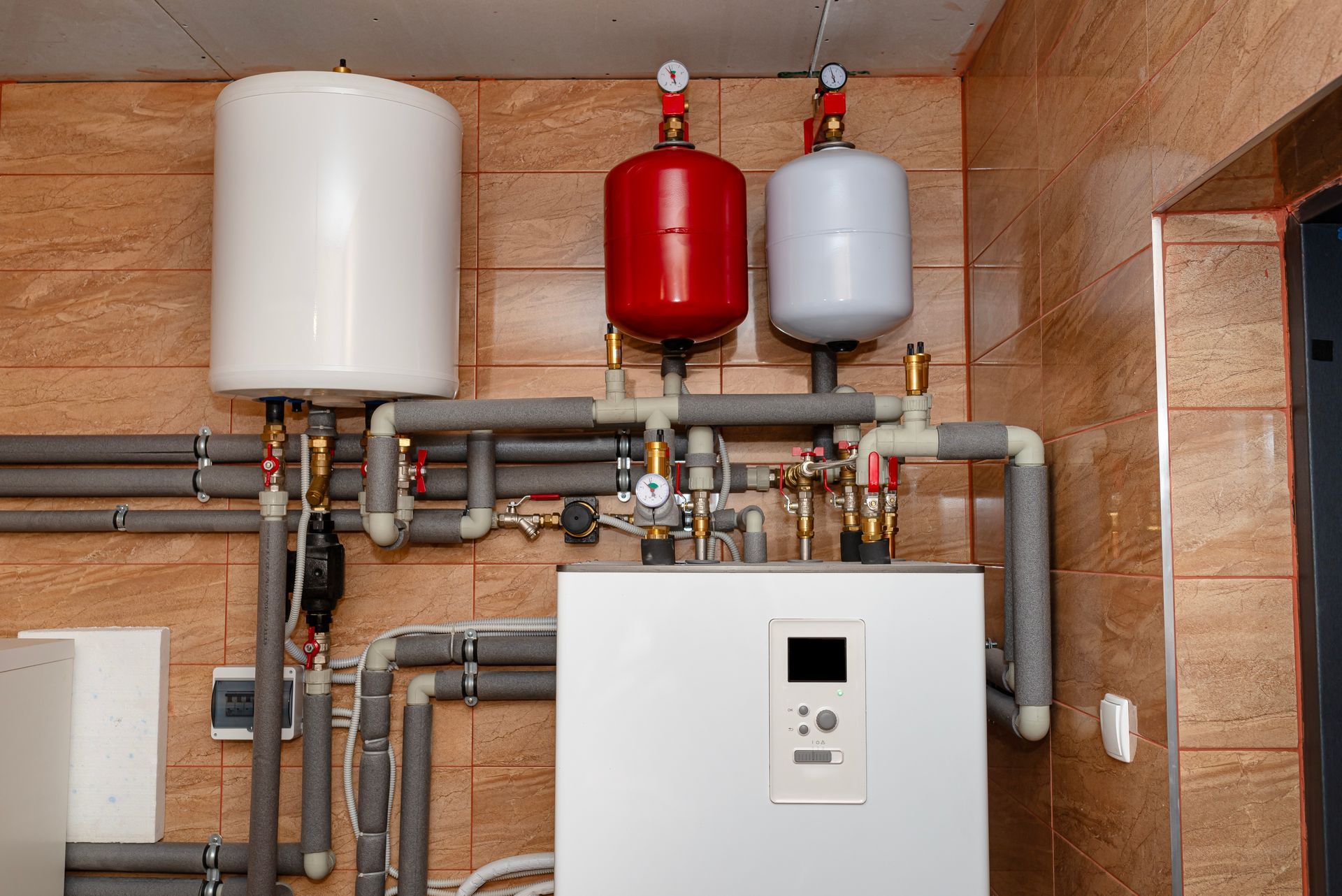Plumbing system with white and red tanks and insulated pipes against a tiled wall.