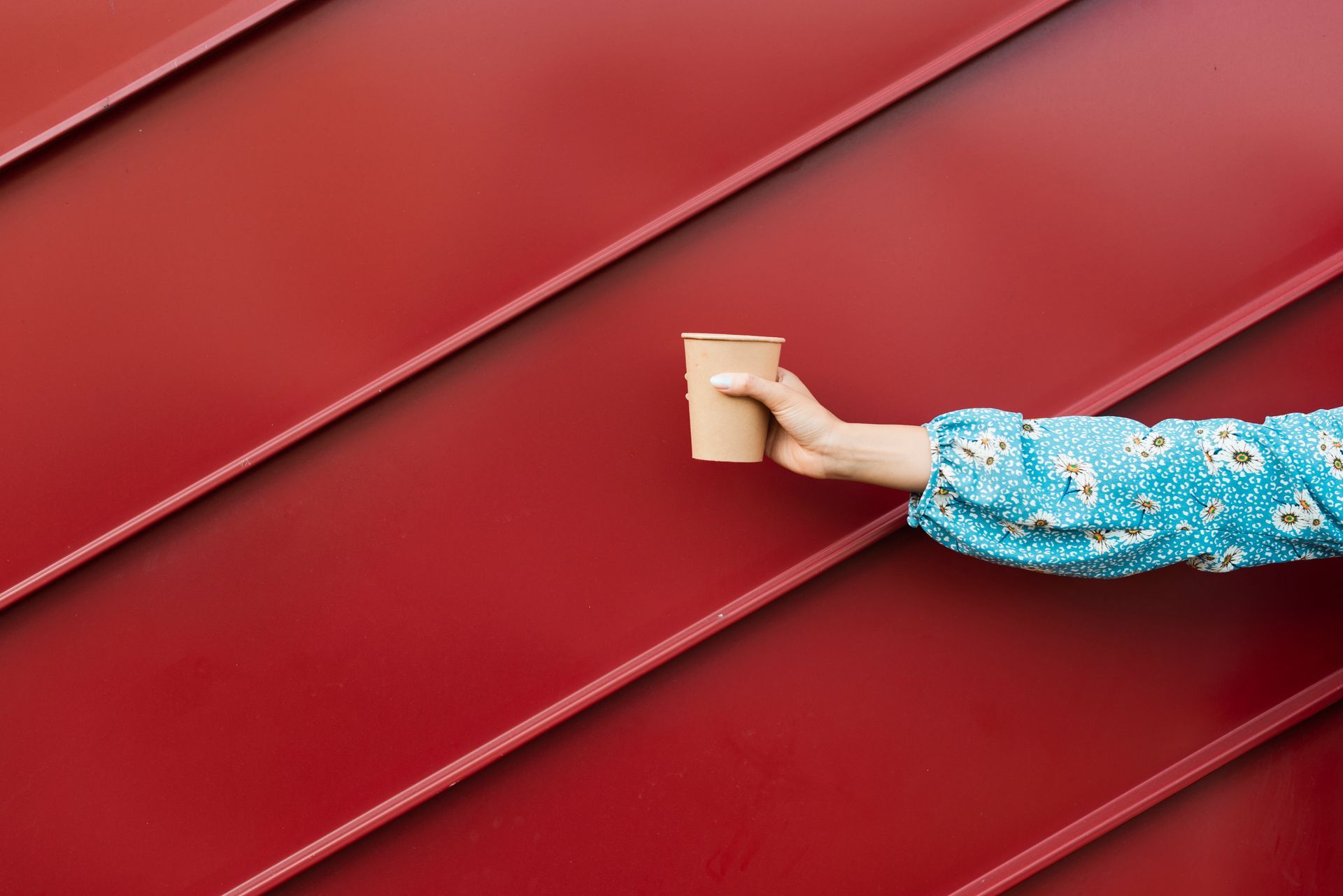 A hand in a blue patterned sleeve holds a tan paper coffee cup against a diagonal, ribbed red background.