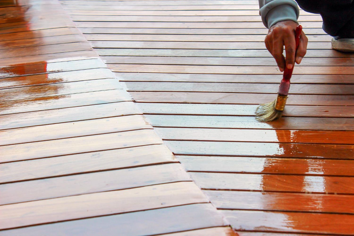 Person applying sealant to a wooden deck with a brush. Half of the deck is sealed, showing a shiny, wet finish.