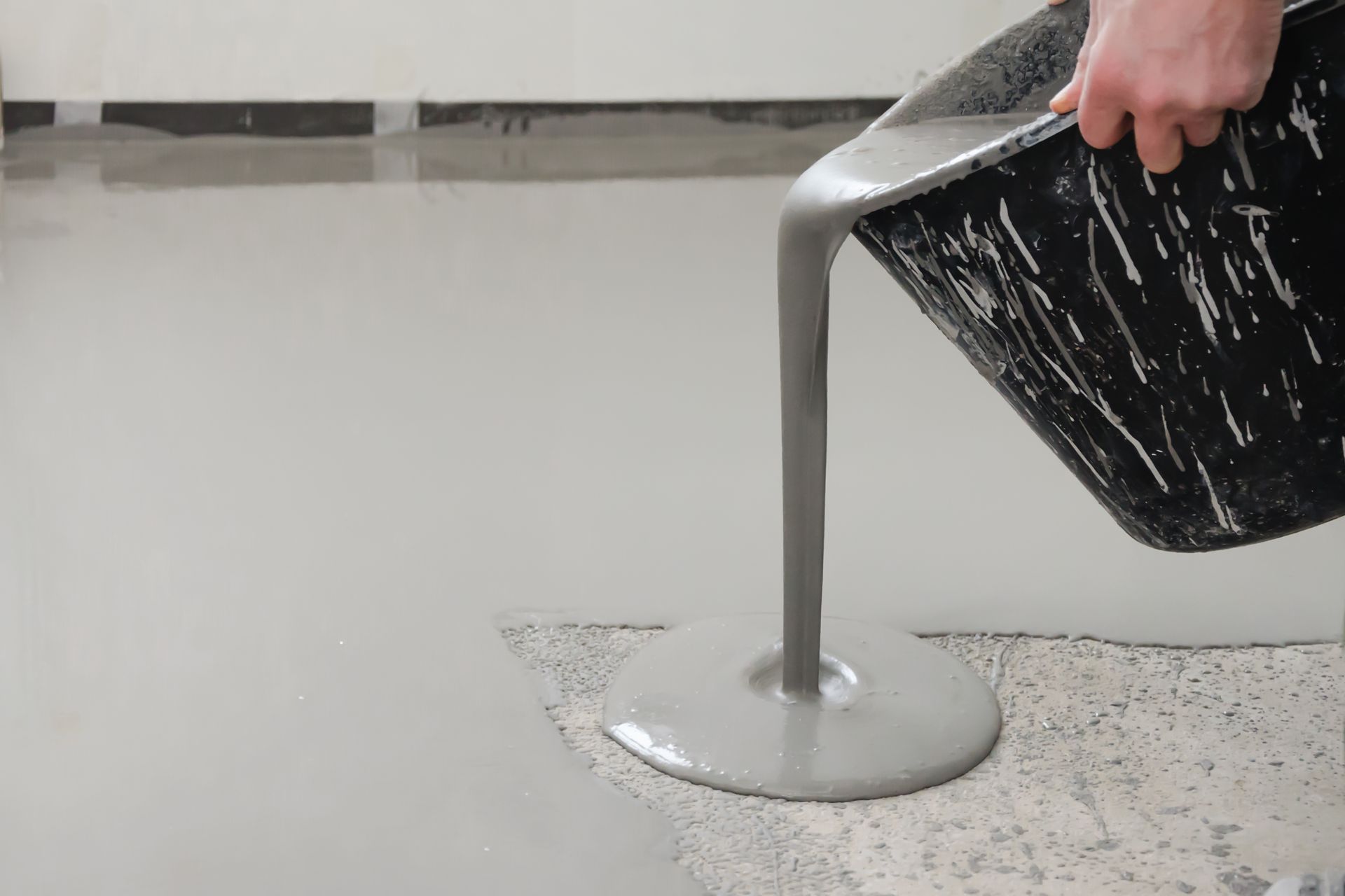 Person pouring gray liquid concrete from a black bucket onto a gray floor.