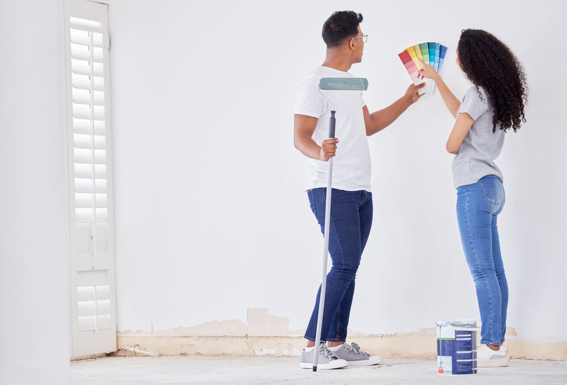 Couple selecting paint colors, man holds roller. White room, paint swatches, jeans, casual clothes.