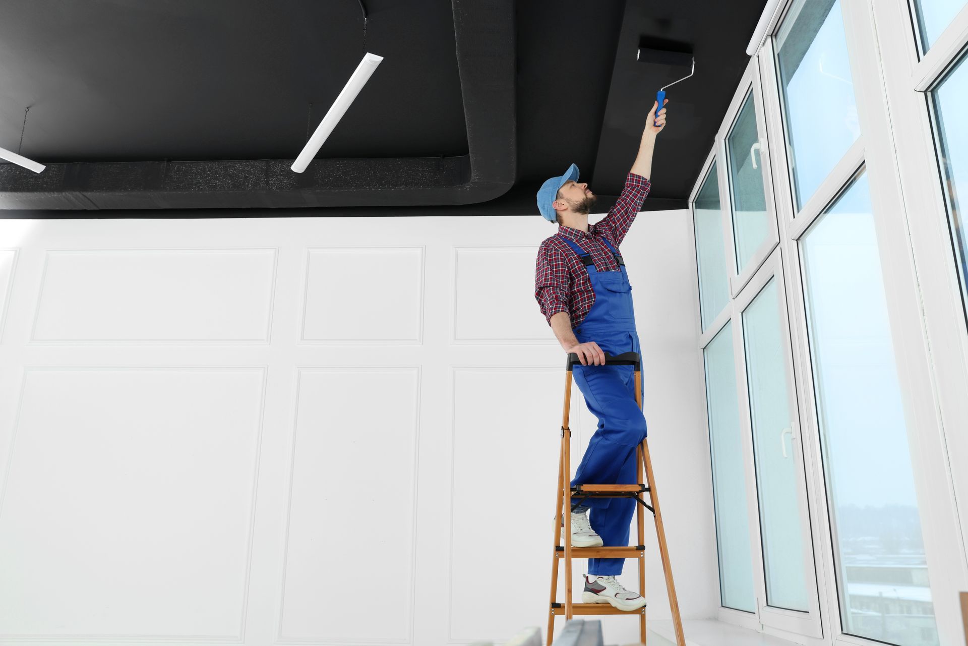 A man is standing on a ladder painting the ceiling with a roller.