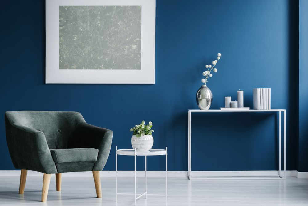 A living room with blue walls , a chair , a table and a picture on the wall.