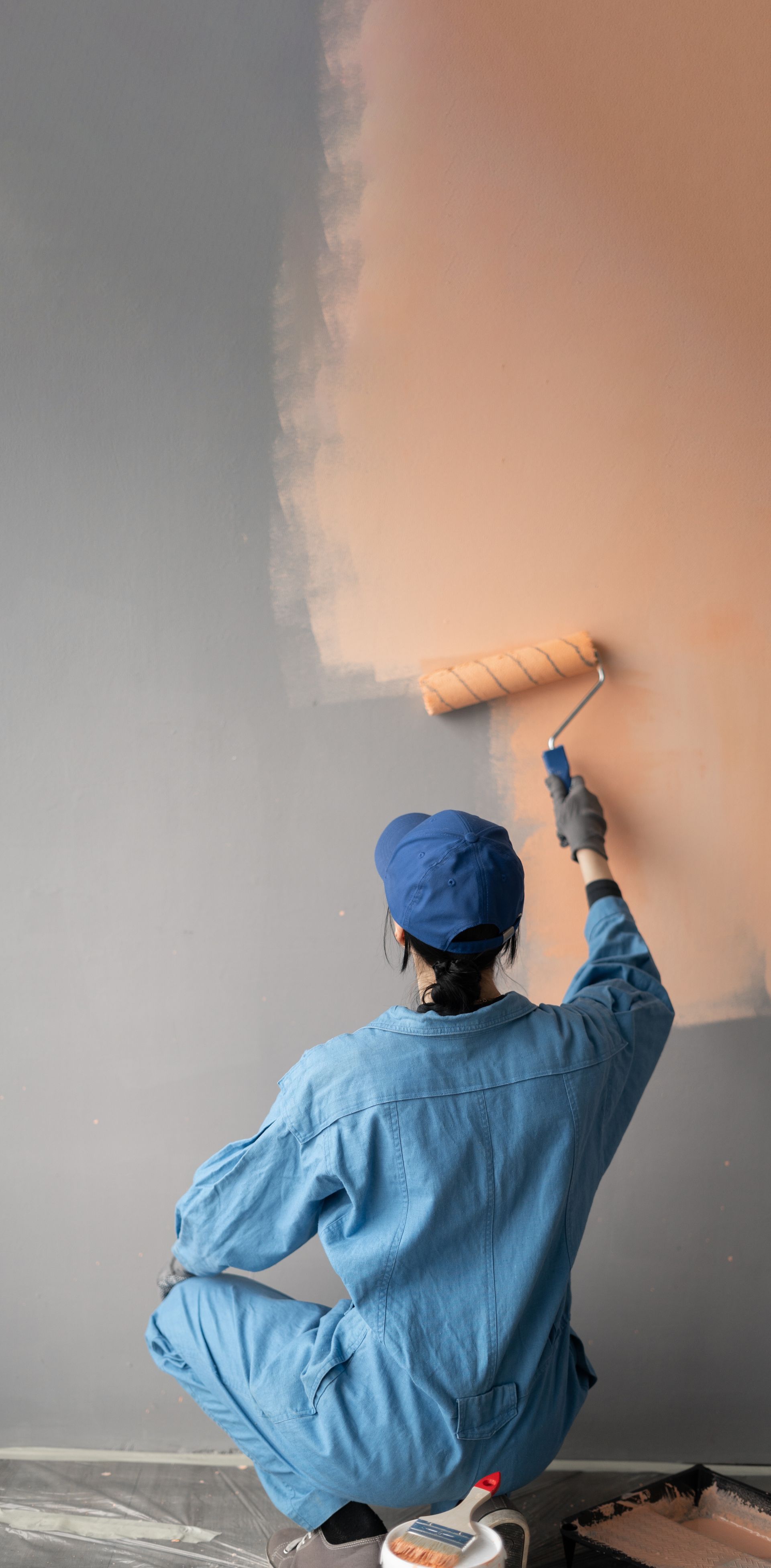 A woman is kneeling down and painting a wall with a paint roller.
