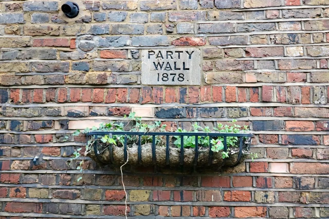 Brick wall with a plaque reading “PARTY WALL 1878” and a small flower planter beneath it