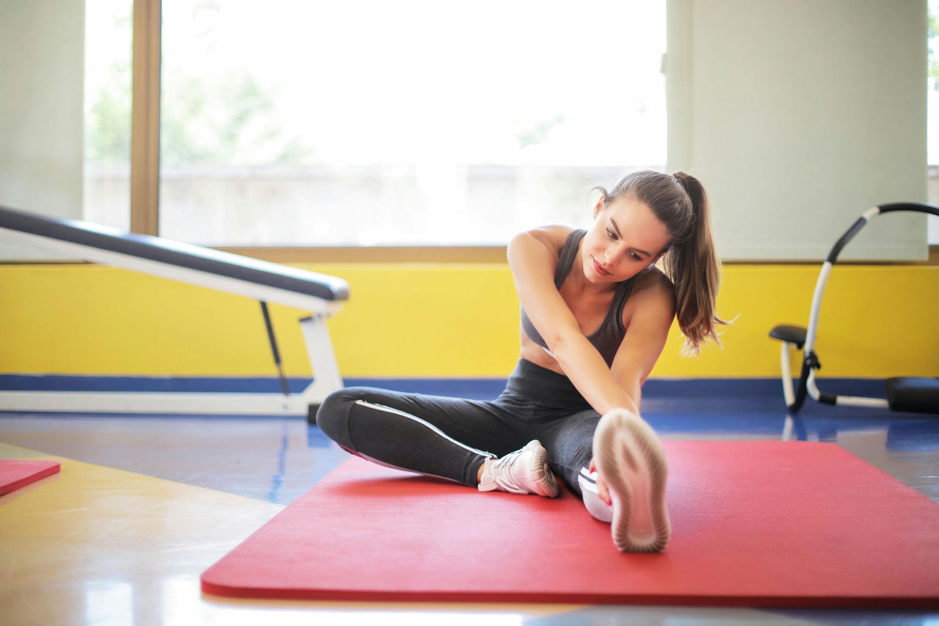 A woman is sitting on a yoga mat in a gym stretching her legs.