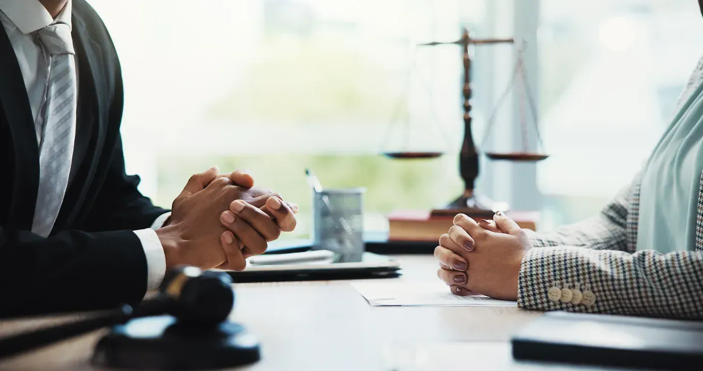 Two people in professional attire seated at a table, hands clasped. A gavel and scales of justice are in the background.