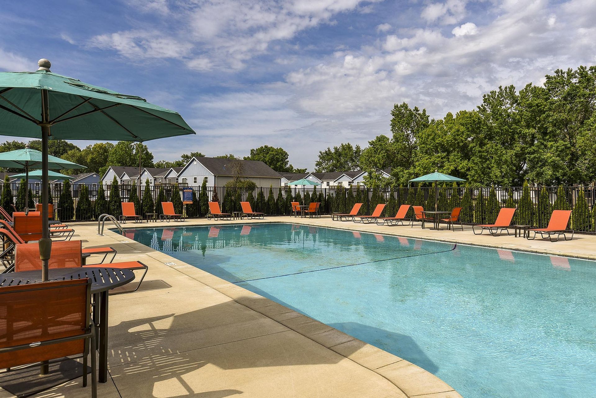 Outdoor community pool with orange loungers, teal umbrellas, and a fenced perimeter.