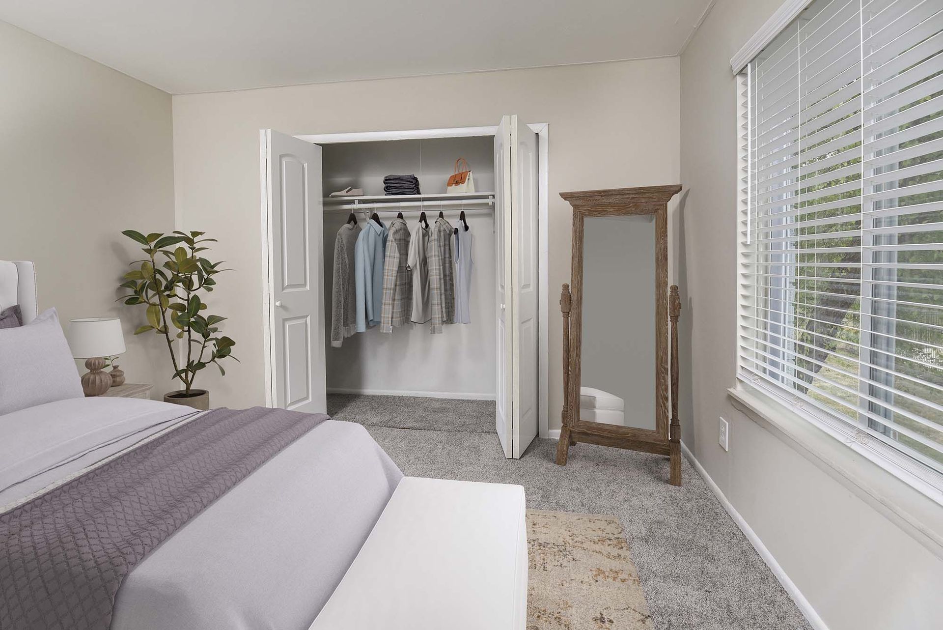 Bedroom with an open closet of hanging clothes, a full-length mirror, blinds on the window, and a bed.