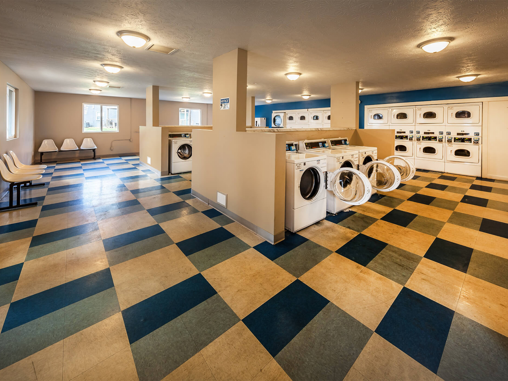 Public laundry room with front-loading washers and dryers, seating area, and a checkered floor.