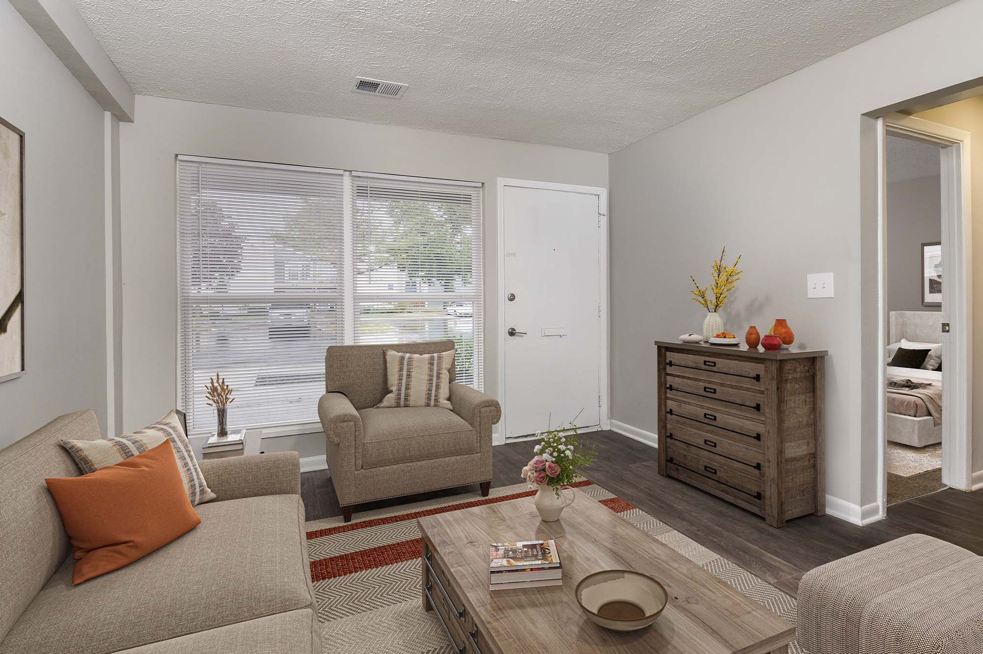 Living room in an apartment with beige sofa and chair, coffee table, and a dresser near a doorway.