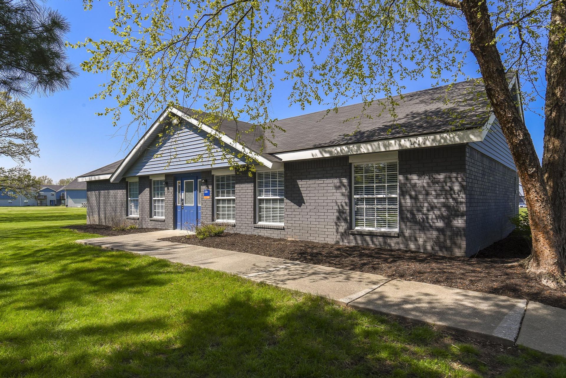 Exterior view of a gray brick apartment building with a blue front door and green lawn.