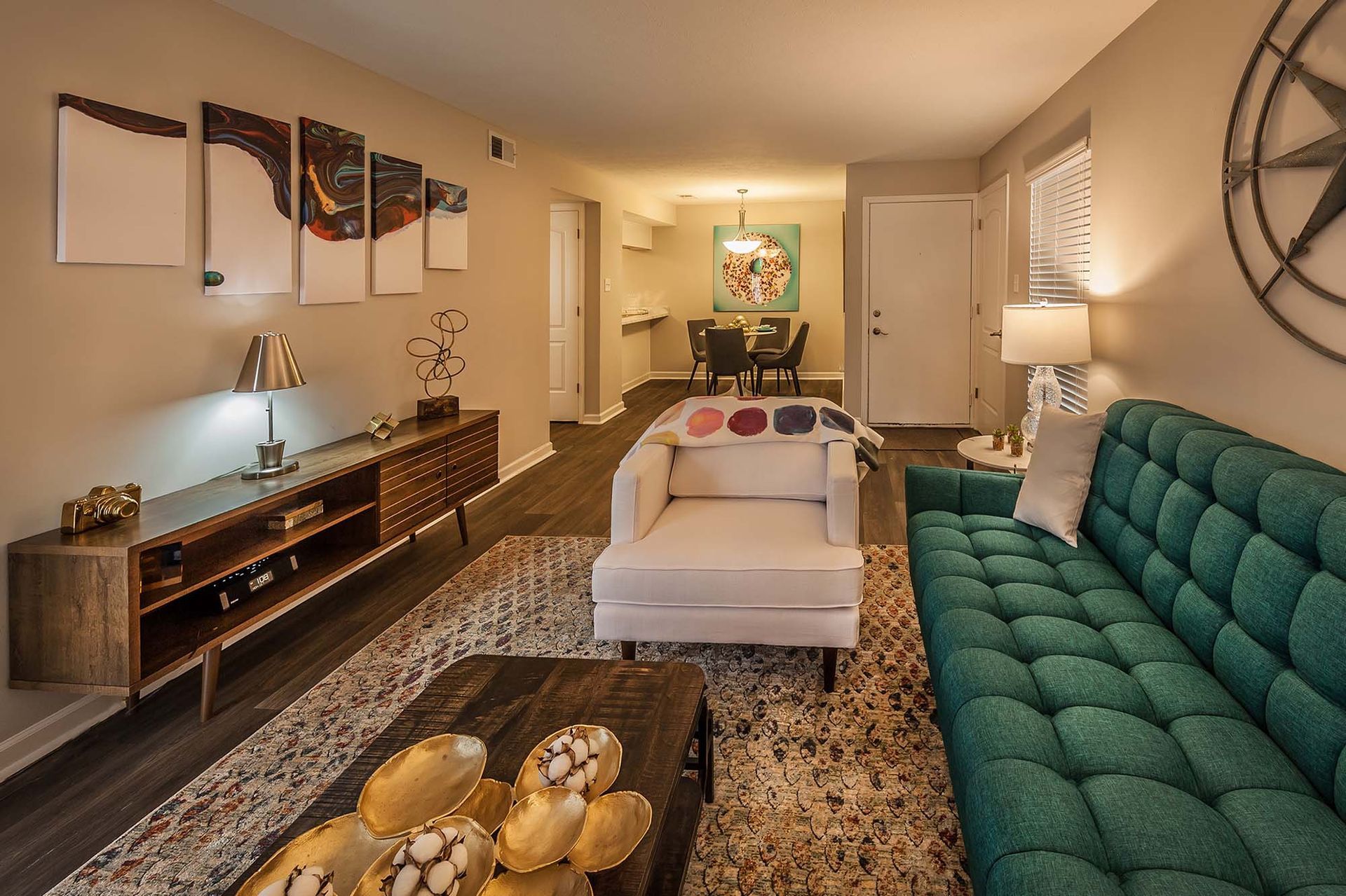 Living room in an apartment with a teal sofa, white chair, and a dining area in the background.