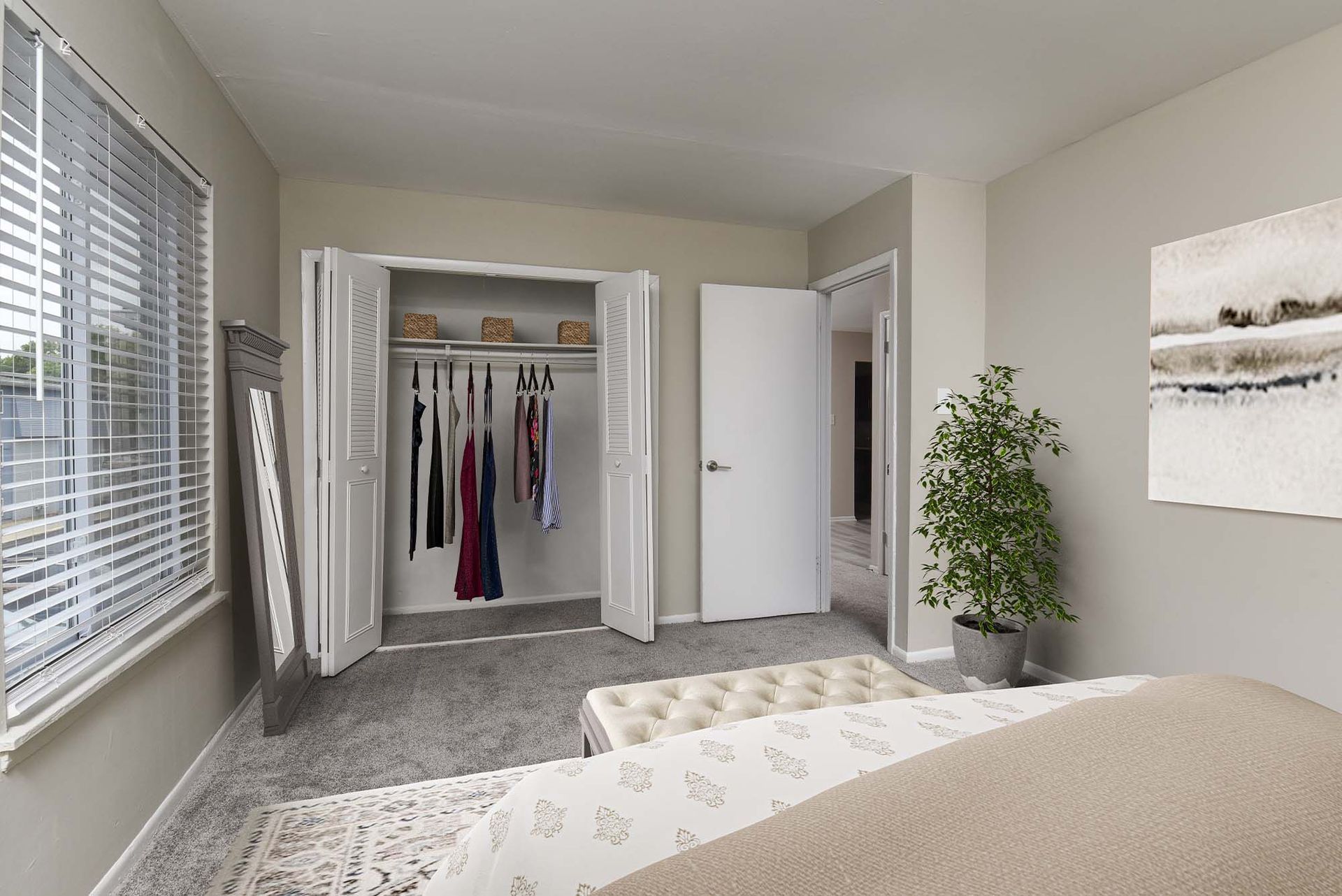 Bedroom interior with an open closet showing hanging clothes, a plant, and a bed.