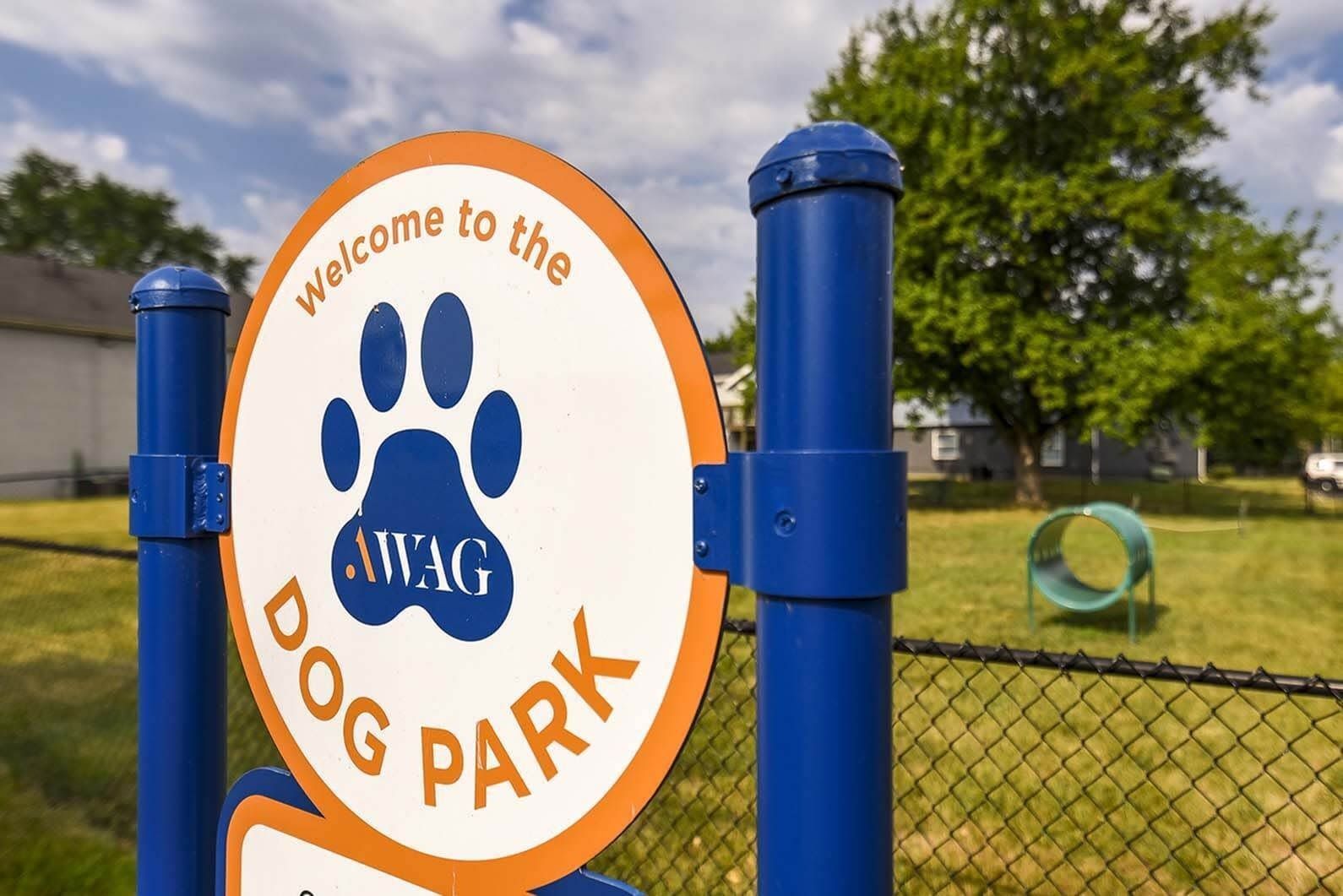 Circular dog park sign with blue paw print and orange border, mounted between blue posts.