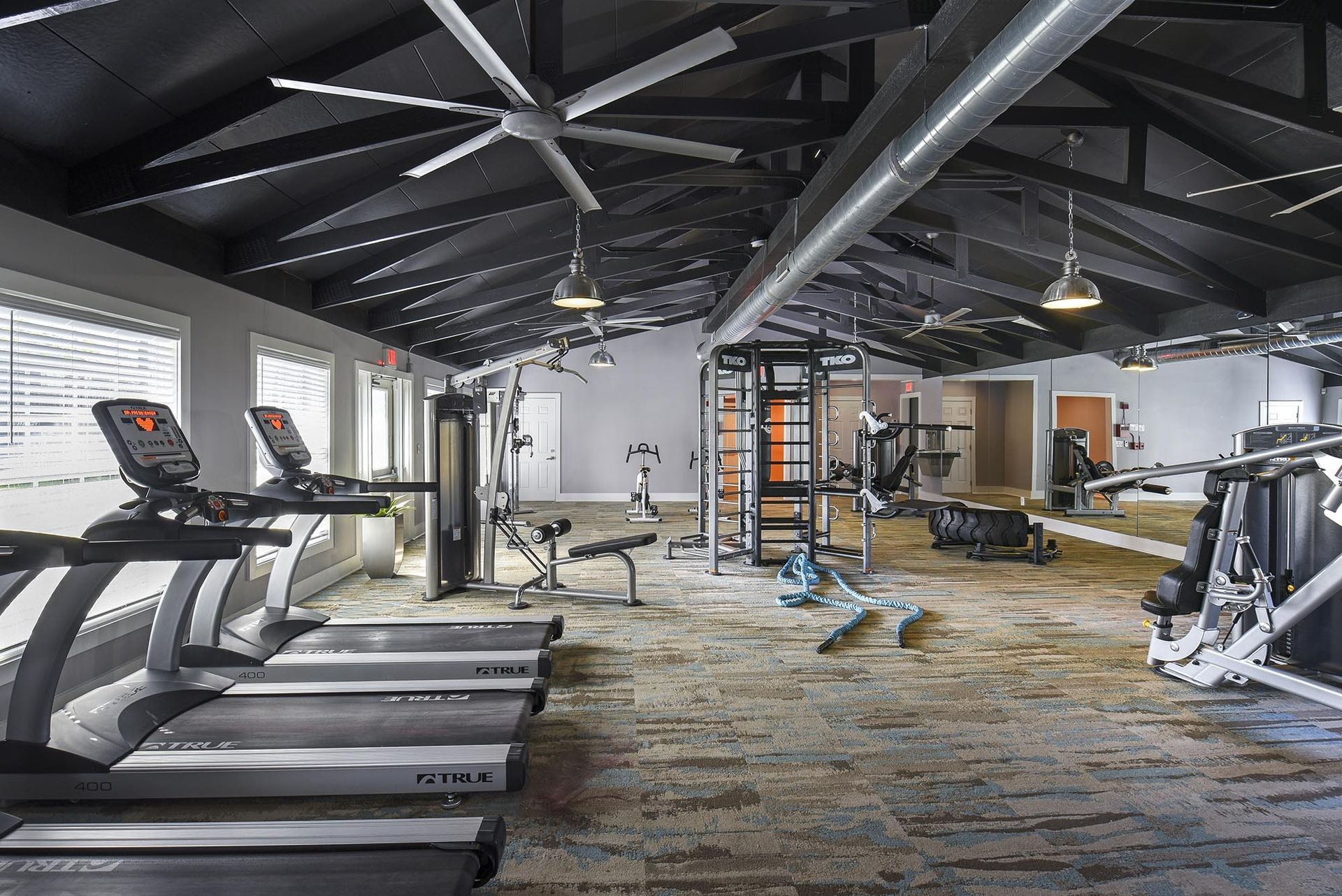 Interior view of a modern apartment gym with treadmills, weight machines, and exposed industrial ceiling beams.