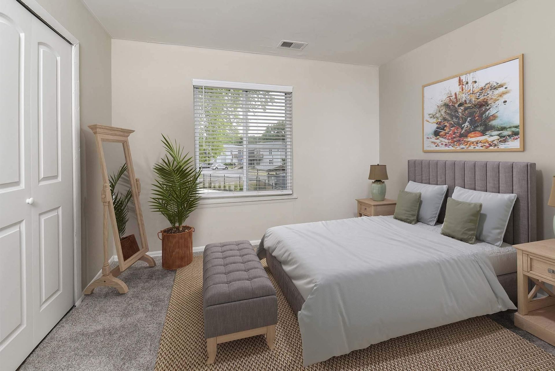 Bedroom in an apartment with a gray upholstered bed, bench, two nightstands, and a window with blinds.