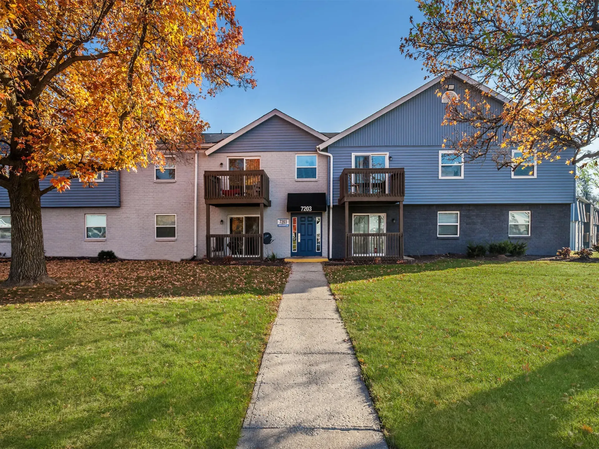 Exterior view of a two-building apartment complex with balconies, central entrance, and autumn trees.