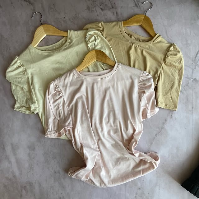 Tres blusas de mujer de colores pastel colgadas en perchas de madera sobre una superficie de mármol.