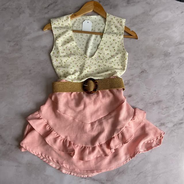 Una blusa floral color crema, una falda con volantes de color rosa y un cinturón color canela con hebilla de madera en una percha.