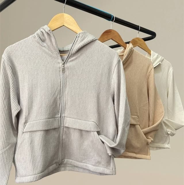 Cuatro chaquetas con capucha y cremallera en gris, tostado y blanco cuelgan de un perchero negro.