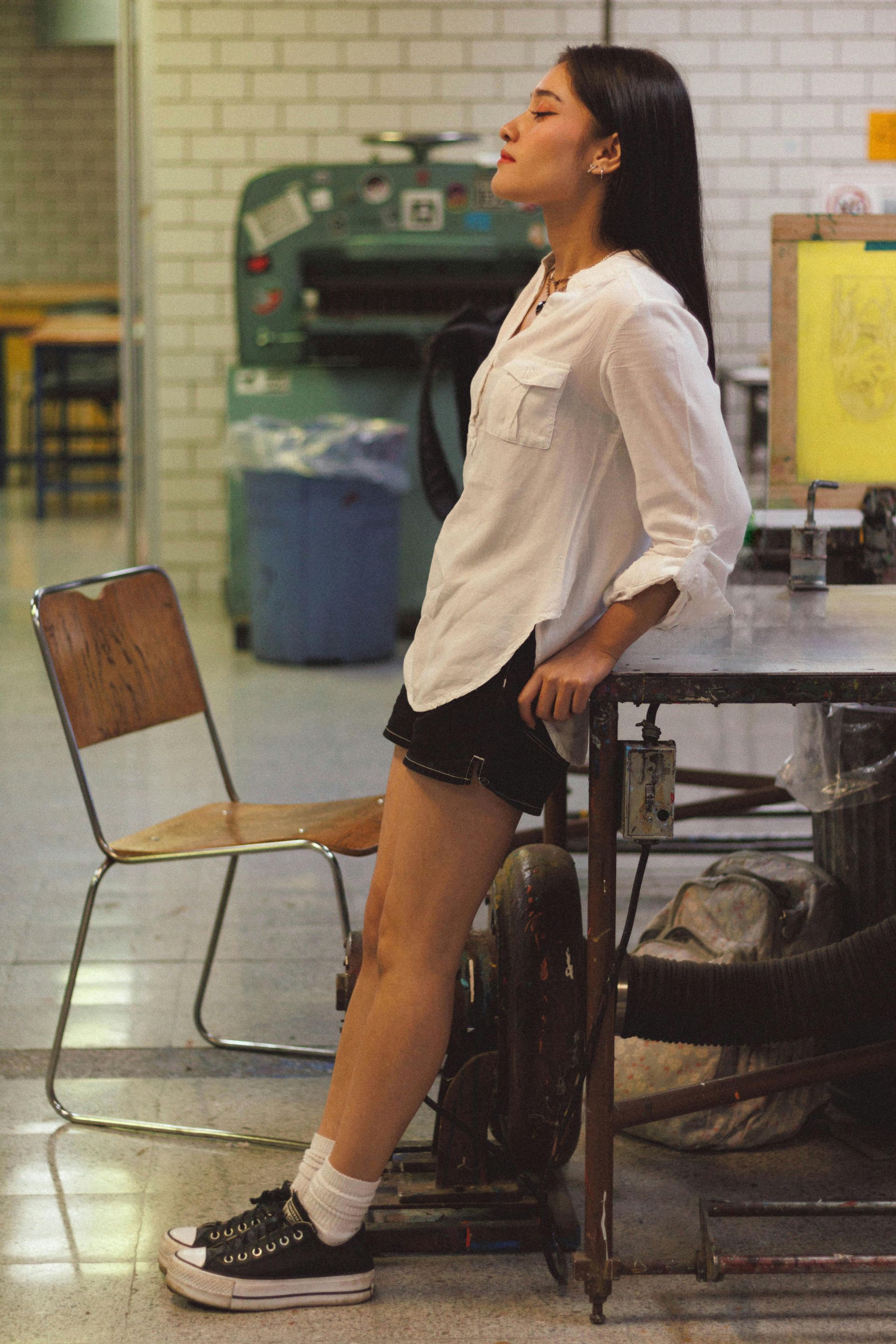 Mujer apoyada en una mesa en un estudio de arte, ojos cerrados, camisa blanca, pantalones cortos negros, zapatillas Converse.