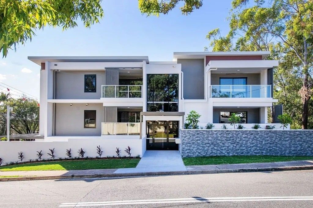 Modern two-story apartment building with balconies, glass entrance, grey and white facade, and stone wall.